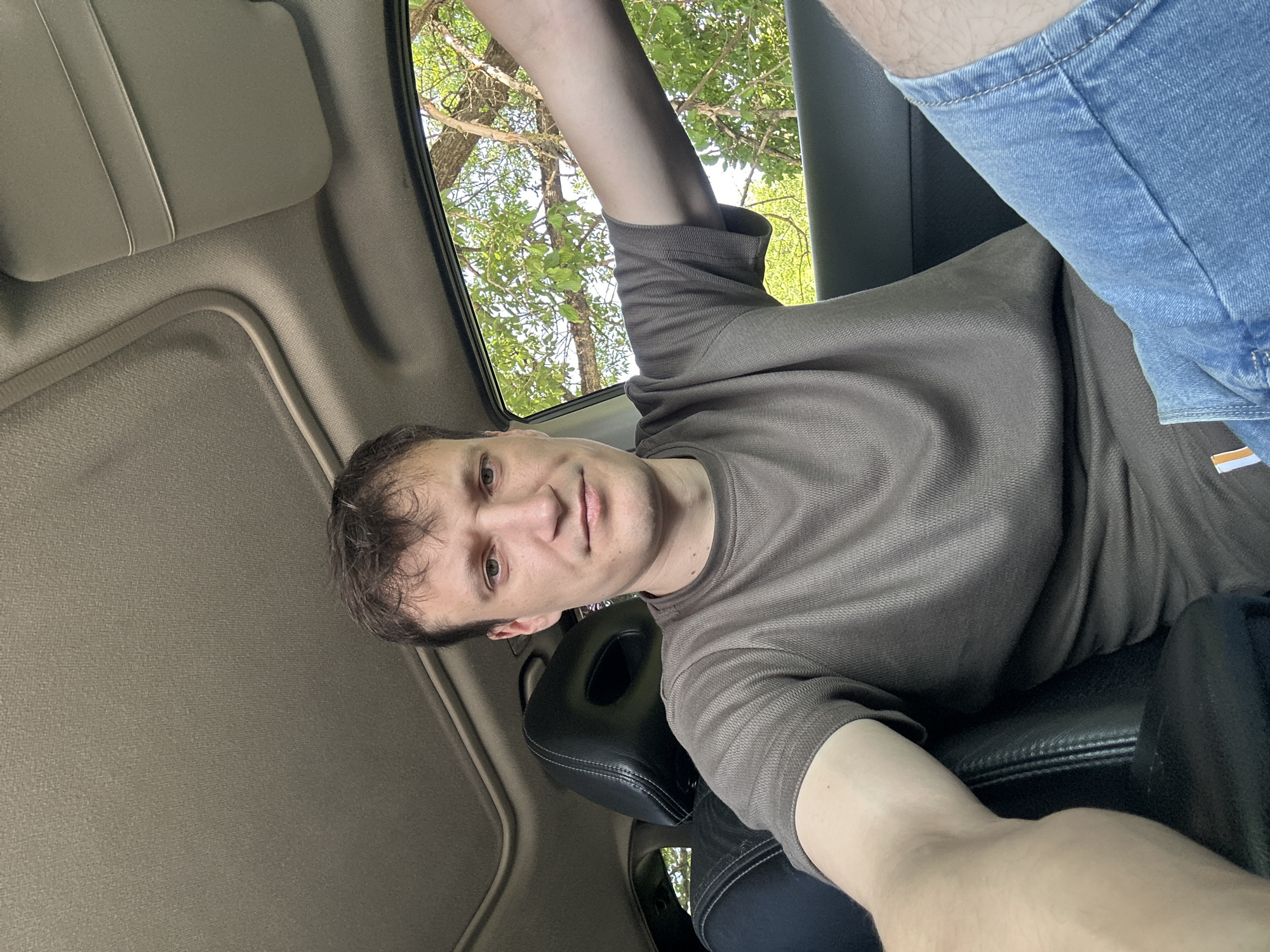 In Makiv, Ukraine, a man with short brown hair and light eyes is seated in the driver's seat of a car. He is wearing a brown t-shirt and blue denim shorts. The car has black leather interior and a sunroof. Through the open window, green trees and foliage are visible, suggesting an outdoor, possibly rural, setting. The man is looking towards the camera, holding the phone to take the selfie.