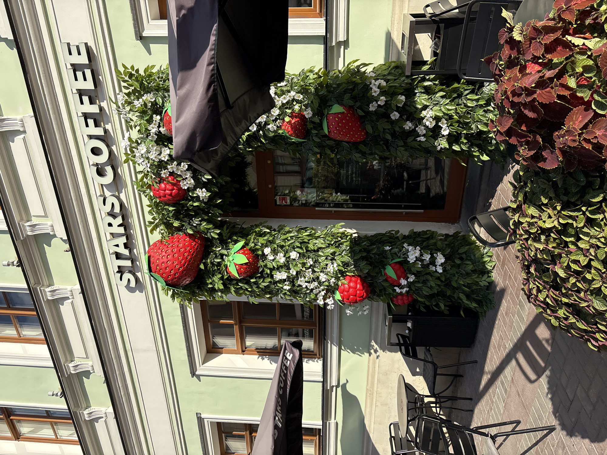 The image shows the entrance to a "STARS COFFEE" establishment decorated with a green leafy arch adorned with large artificial strawberries and white flowers. The building facade is light green with white decorative molding and wooden-framed windows. Outside the entrance, there are outdoor seating areas with black metal tables and chairs, a black trash bin, and a black umbrella with the "STARS COFFEE" logo. The ground is paved with red bricks. The sun casts shadows on the pavement, indicating it is a sunny day. The photograph was taken in Moscow, Russia.