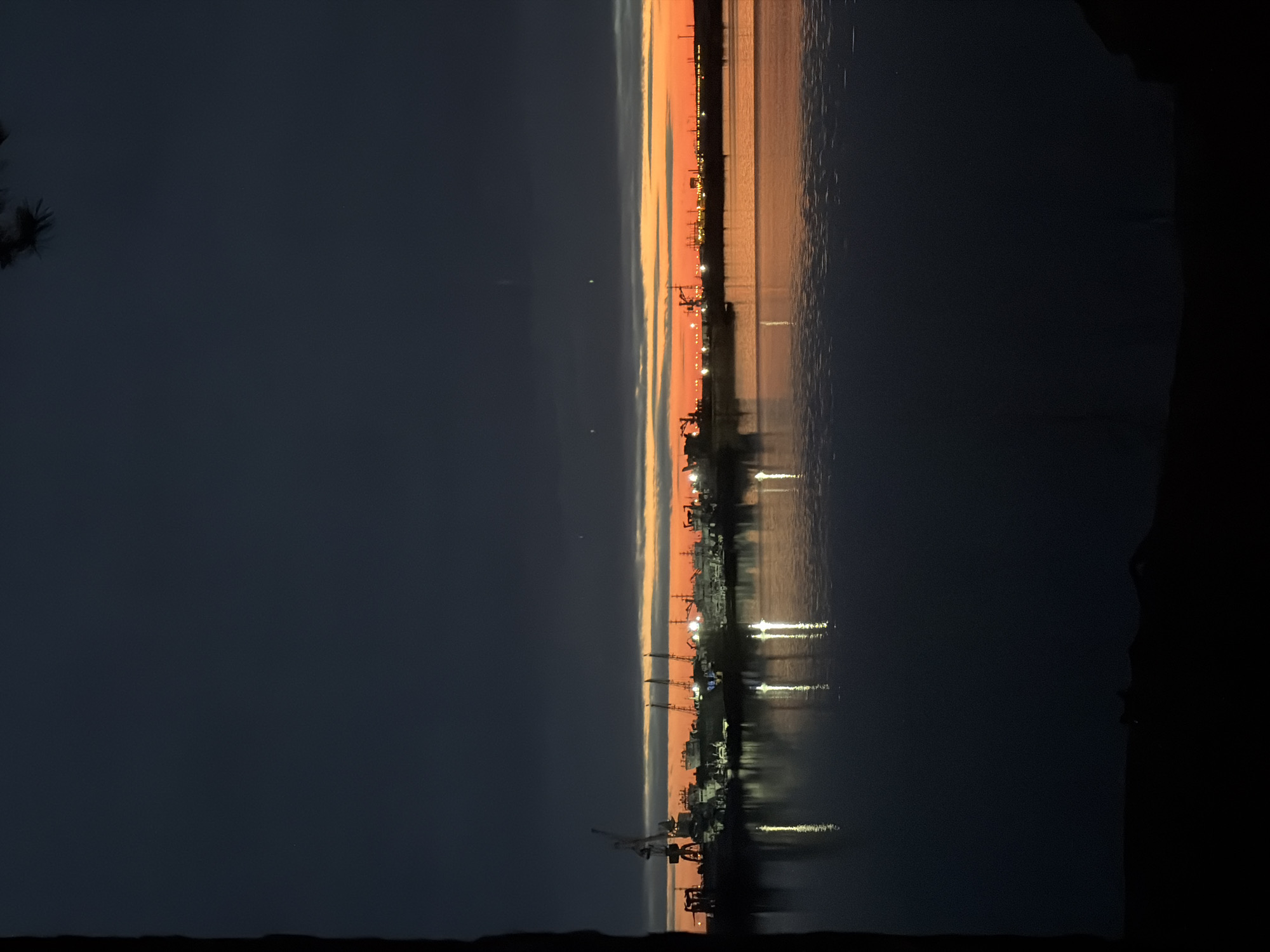 A horizontal photograph captures a tranquil twilight scene over a body of water, likely the Kama River in Chaikovsky, Russia. The sky transitions from a deep, dark blue at the top to a vibrant band of orange and yellow along the horizon, indicating either sunrise or sunset. Wispy clouds are visible in the upper sky, while the lower horizon is dominated by the silhouette of an industrial shoreline.
Along this distant bank, numerous structures resembling cranes and other port machinery are visible, their dark forms contrasting sharply with the bright sky. Small, artificial lights dot the shoreline, some appearing as bright pinpricks, others forming more extensive illuminated areas.
The lower half of the image is occupied by the calm surface of the water, which beautifully reflects the colors of the sky and the twinkling lights from the distant shore, creating elongated, shimmering streaks of light. In the extreme upper left corner, the dark, silhouetted branches of a tree or bush are slightly visible, suggesting the vantage point of the photographer. No people or visible text are present in the image. The overall atmosphere is serene, highlighting the blend of natural beauty and industrial presence at the edge of the water.
