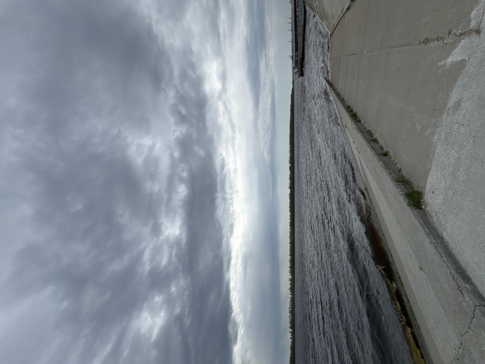 The image shows a concrete embankment or dam on a cloudy day. The water is dark and choppy. Some greenery grows along the edge of the concrete structure. The sky is filled with heavy, gray clouds. The photo was taken in Nizhnevartovsk, Russia.