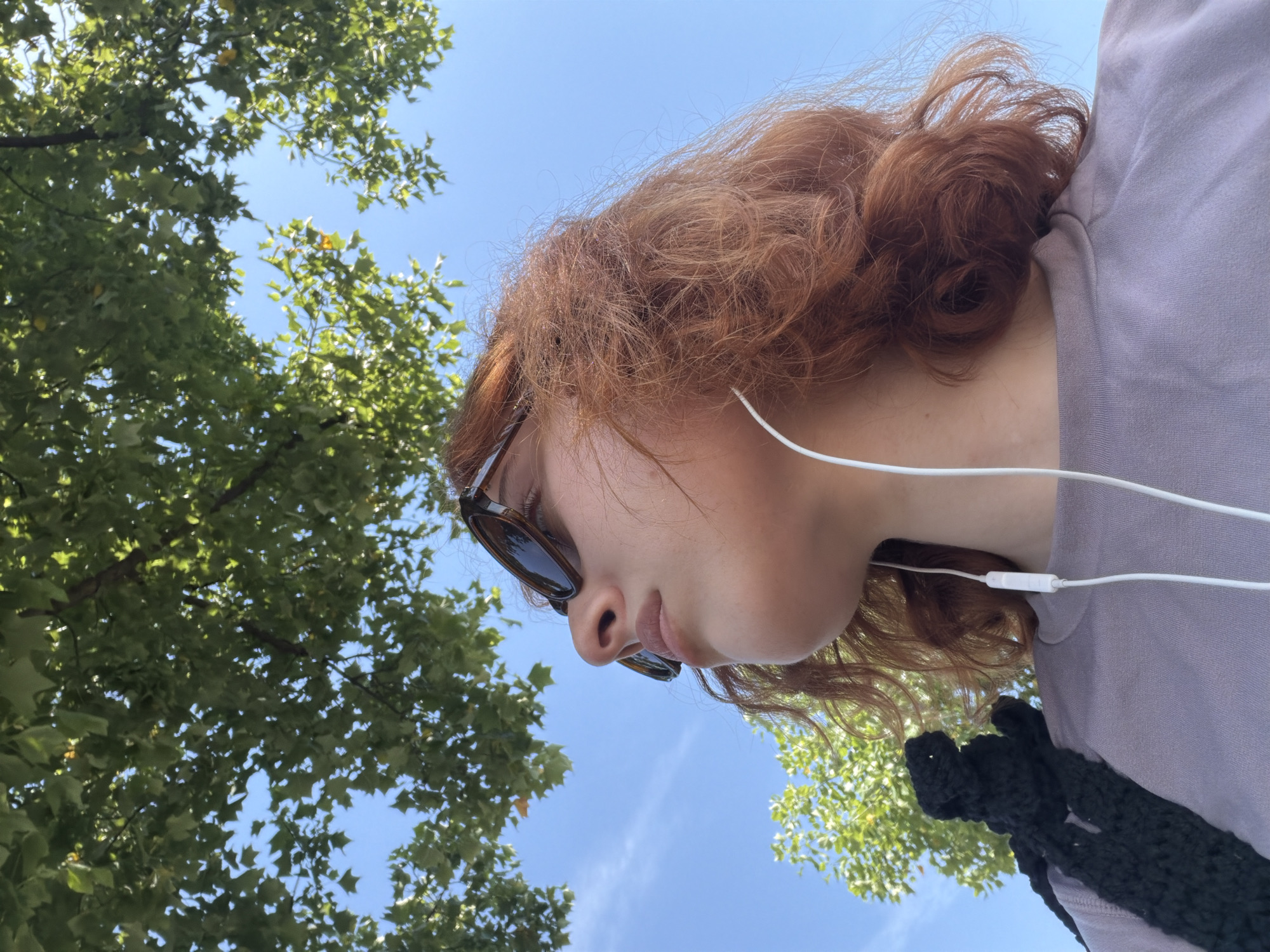 A low-angle, outdoor, close-up shot captures a person from the chest up, looking towards the sky. The person has reddish-brown, wavy hair and is wearing sunglasses and white earbuds. They have a dark, woven bag slung over their left shoulder. The background consists of a clear blue sky and green foliage from trees. The overall lighting suggests it is daytime, with the sun illuminating the scene from above. The image does not contain any discernible elements that would allow for the identification of the city of Strasbourg, France.