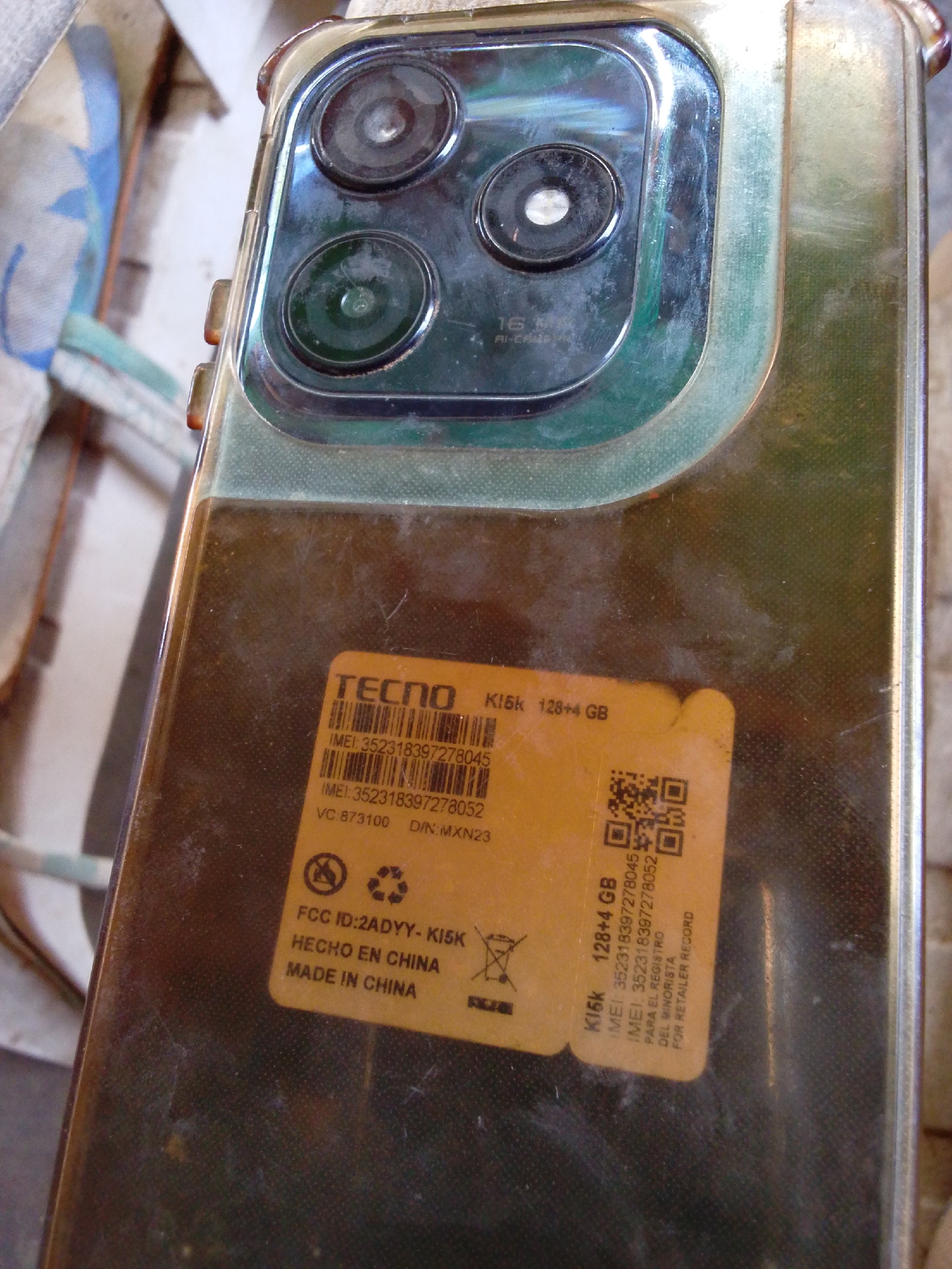 The media file displays a close-up, slightly angled view of the back of a "TECNO K15k" smartphone, encased in a transparent or translucent protective cover. The phone features a prominent camera module with three lenses arranged vertically on a dark, rectangular bump. Faint text near the cameras reads "16 MP AI-CAM".
A yellow-orange sticker is affixed to the lower part of the phone's back, providing various product details. Visible text on the sticker includes "TECNO K15k 128+4 GB", two IMEI numbers ("352318397278045" and "352318397278052"), "VC:873100 D/N:MXN23", "FCC ID:2ADYY-K15K", "HECHO EN CHINA", and "MADE IN CHINA". Additionally, it states "PARA EL REGISTRO DEL MINORISTA" and "FOR RETAILER RECORD", indicating it's an informational label for retail purposes. A QR code is also present on the sticker.
The phone and its case appear somewhat dusty or scratched, suggesting use. The background is out of focus, showing indistinct light-colored fabric or soft material, possibly an upholstered surface, indicating an indoor setting. The lighting suggests daytime, but specific time or weather is not discernible.