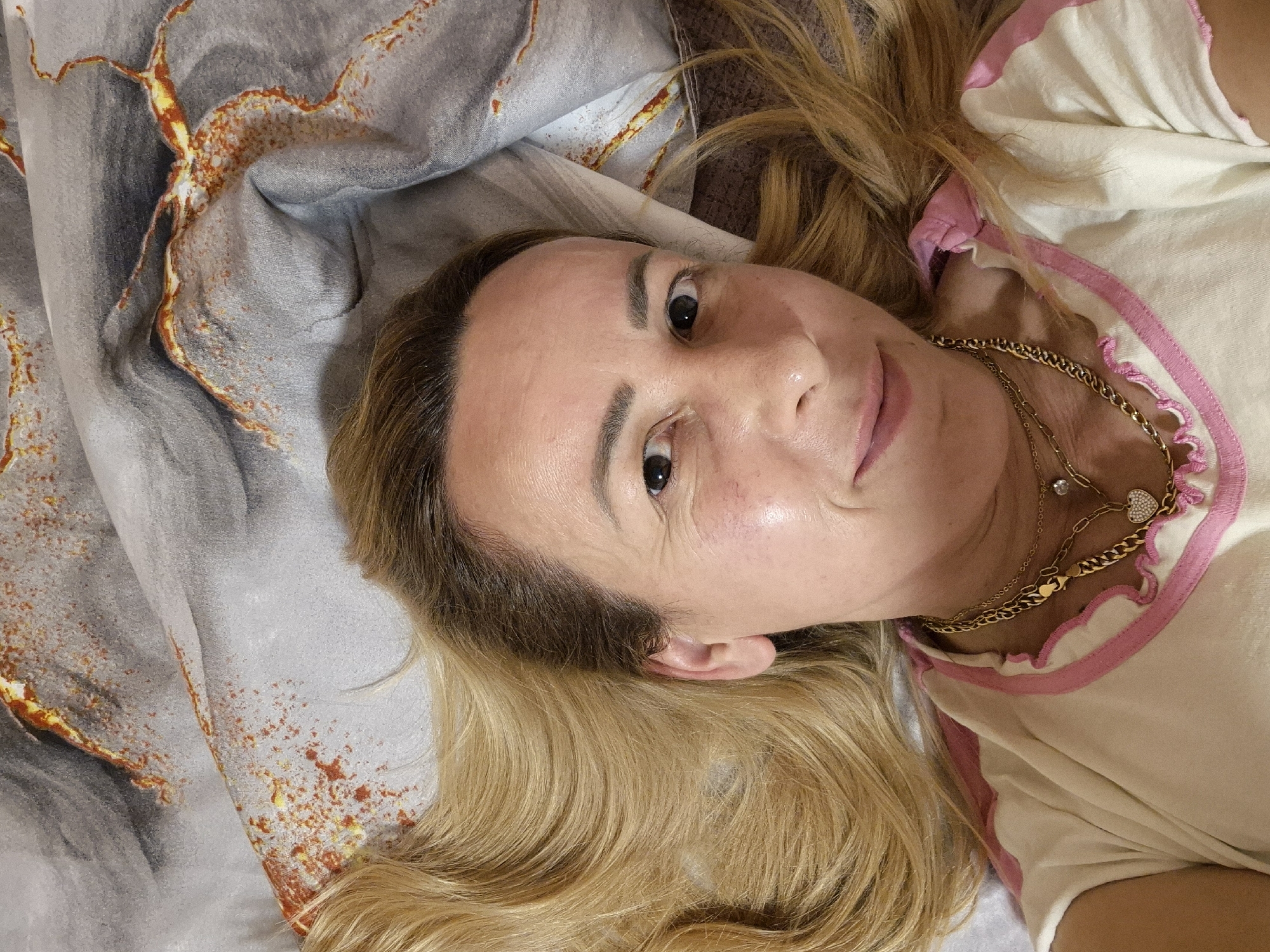 A woman with blonde hair lies on her back on a bed. She is wearing a white shirt with pink trim. The background appears to be a duvet with a marbled pattern of gray, white, and orange. She is wearing a gold necklace with a heart-shaped pendant. Her eyes are open and she is looking at the camera. The lighting is bright.