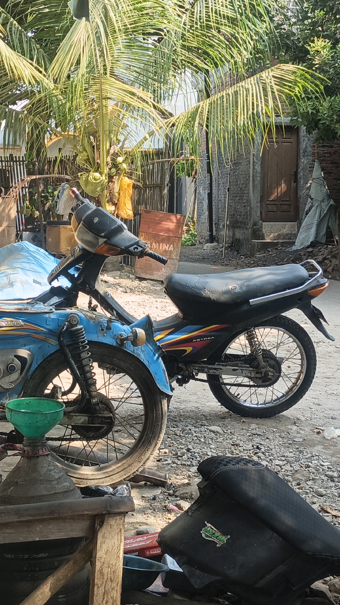 The image depicts an outdoor scene with two motorcycles partially visible. In the foreground, a blue motorcycle is positioned on the left, with its front wheel and part of the engine showing. A green funnel is placed on a conical object next to it. To the right, a black motorcycle is parked, with its seat clearly visible, showing signs of wear. The background features a coconut tree with fruits and leaves, a wooden fence, a building with a wooden door, and some rusty barrels. The ground is covered with gravel and dirt. The scene appears to be in a residential or rural area, possibly in Bulungcangkring, Indonesia, given the tropical vegetation. The lighting suggests it is daytime, with sunlight casting shadows. There are no people visible in the image.