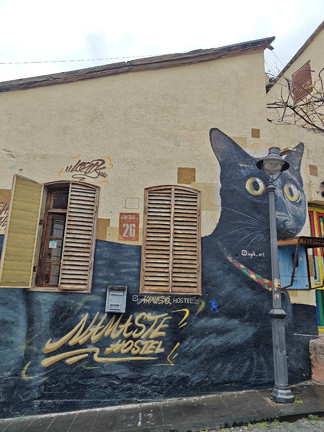 The image captures a vibrant building facade adorned with extensive street art, likely depicting a hostel entrance. The upper portion of the building is painted in a light beige stucco, transitioning to a dark blue-grey mural covering the lower half.
A prominent feature is a large mural of a black cat with wide, bright yellow eyes, depicted on the right side of the wall, seemingly peering around a tall, black streetlamp. The cat wears a colorful, braided collar. To the left, two windows with wooden louvered shutters interrupt the mural. The left window has one shutter open and the other partially open, while the right window's shutters are closed. Between these windows, a small grey utility box or mail slot is mounted on the wall.
Above the windows, a reddish-brown plaque displays the number "26." Faint, brown and orange graffiti, possibly spelling "welcome," is visible above the left window. Below the mail slot, large, stylized golden letters spell out "NAMASTE HOSTEL." Additional text includes Instagram handles: "@NAMASR_HOSTEL" near the mail slot and "@logik_art" near the cat's head, likely crediting the artist.
On the far right, a colorful structure, possibly a painted telephone booth, is partially visible, with the word "TELEPHON" faintly discernible. The streetlamp in the foreground has a grey lantern. The ground below is a dark, wet-looking sidewalk or pavement, with some greenery at its edge. The sky overhead is overcast, indicating a cloudy daylight scene. The mural transforms the building into a distinctive and eye-catching landmark.