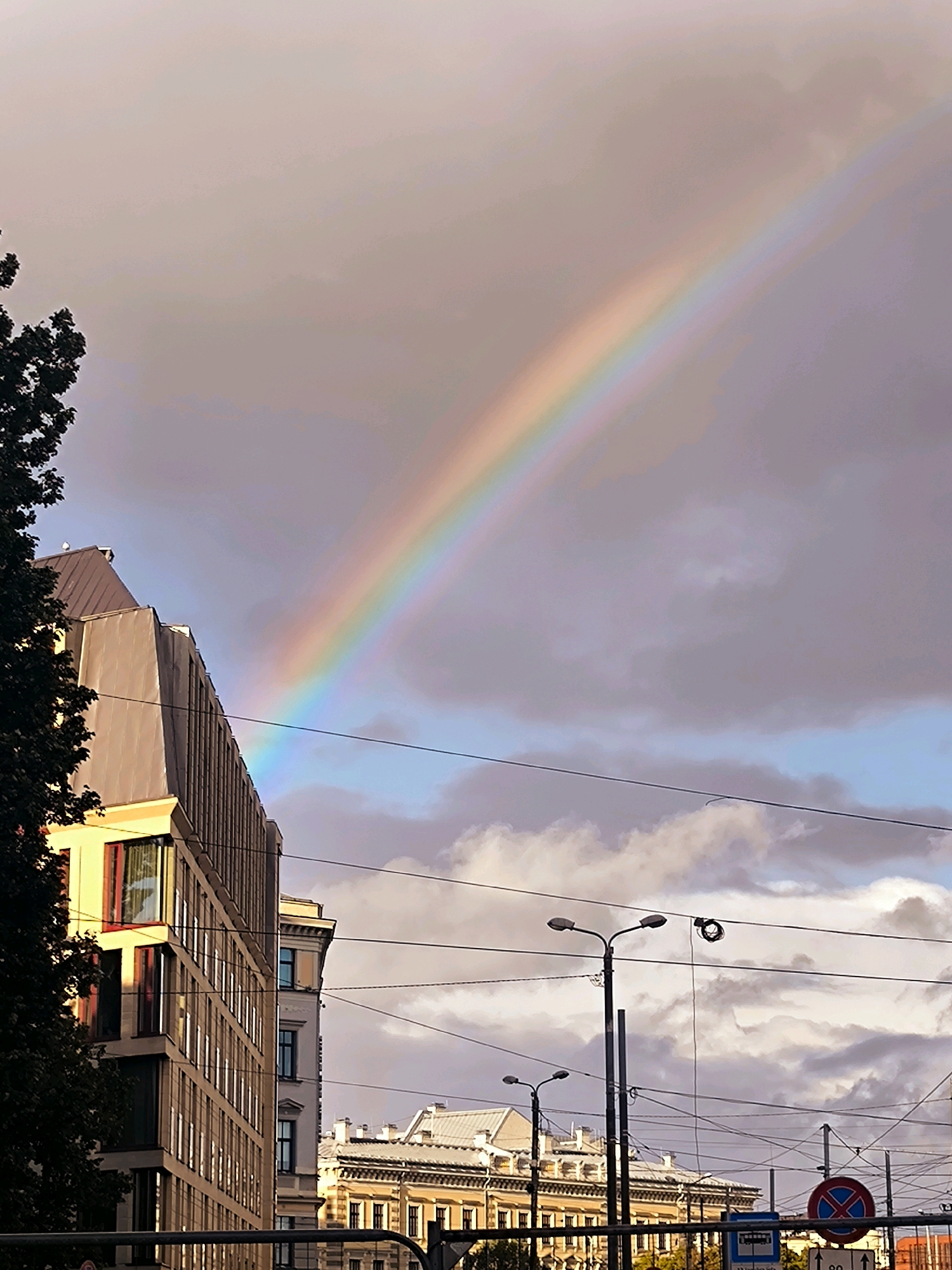 The image captures a city scene in Riga, Latvia, featuring a prominent rainbow arching across a cloudy sky. In the foreground, a modern, multi-story building with a light-colored facade and numerous windows is visible on the left. To the right of this building, older, ornate architecture with cream-colored walls and dark roofs can be seen. The sky is a mix of gray clouds and patches of blue, with the rainbow forming a vibrant arc of colors. Several utility poles with numerous wires crisscross the frame, and streetlights are present. A dark-leafed tree is partially visible on the far left. In the lower right corner, a circular sign with a red border and a diagonal red line over a blue background, likely indicating a no-parking or restricted zone, is visible, along with a traffic sign indicating speed limits. No people are clearly depicted in the scene.