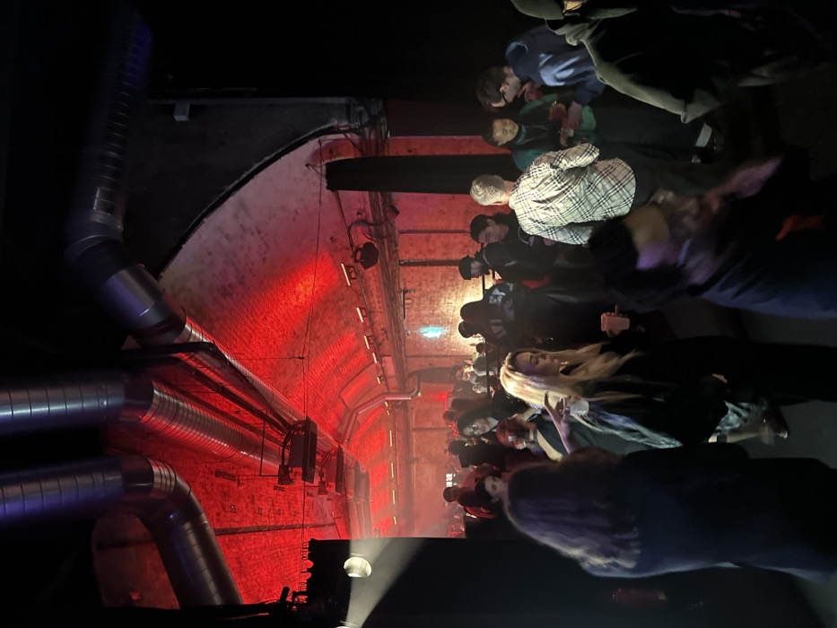 The image displays an indoor gathering within a large, arched brick tunnel, characteristic of a repurposed railway arch venue in London, United Kingdom. The brick ceiling is bathed in a strong red ambient light, highlighting industrial metallic ventilation ducts and visible lighting fixtures. Approximately 20-30 individuals are present, standing and mingling in the dimly lit space. In the foreground, a person with dark hair is seen from behind, beside a blonde-haired woman in a dark jacket holding a light-colored cup. A blurred individual with dark curly hair and glasses is partially visible in the bottom right. In the midground, a man in a plaid shirt is seen from behind, surrounded by other attendees, some holding beverages. The background shows additional individuals dispersed further into the archway, silhouetted against the red illumination. The scene depicts a social event or club night.