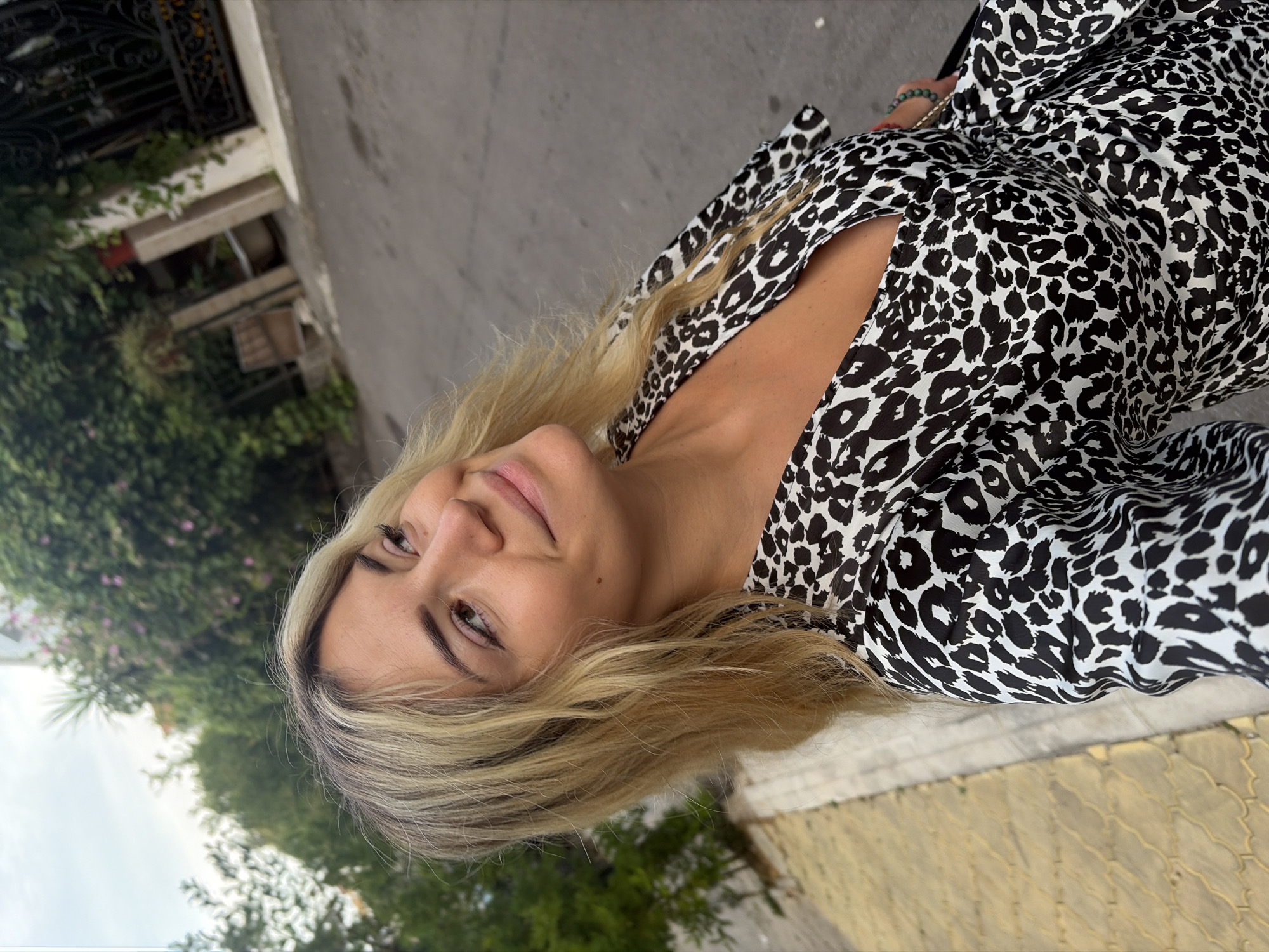 A young woman with blonde hair and a leopard-print dress is standing on a street in Budva, Montenegro. She is looking to her right and smiling. The background is a residential area with trees and buildings. The street is paved, and there is a yellow-tiled sidewalk on the left side of the image. The sky is cloudy.