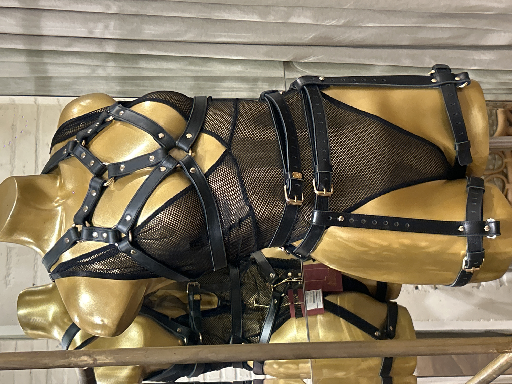 A black mesh bodysuit with a leather harness is displayed on a gold mannequin in a retail setting. The harness features multiple straps, buckles, and metal rings, crisscrossing the chest and torso. Additional straps extend down the legs, attaching to a belt around the thighs. The mannequin is positioned in front of a grey curtain, with a reflection of another similar mannequin visible to the left. The overall lighting is soft and diffused. The location is specified as North Kuta, Indonesia.