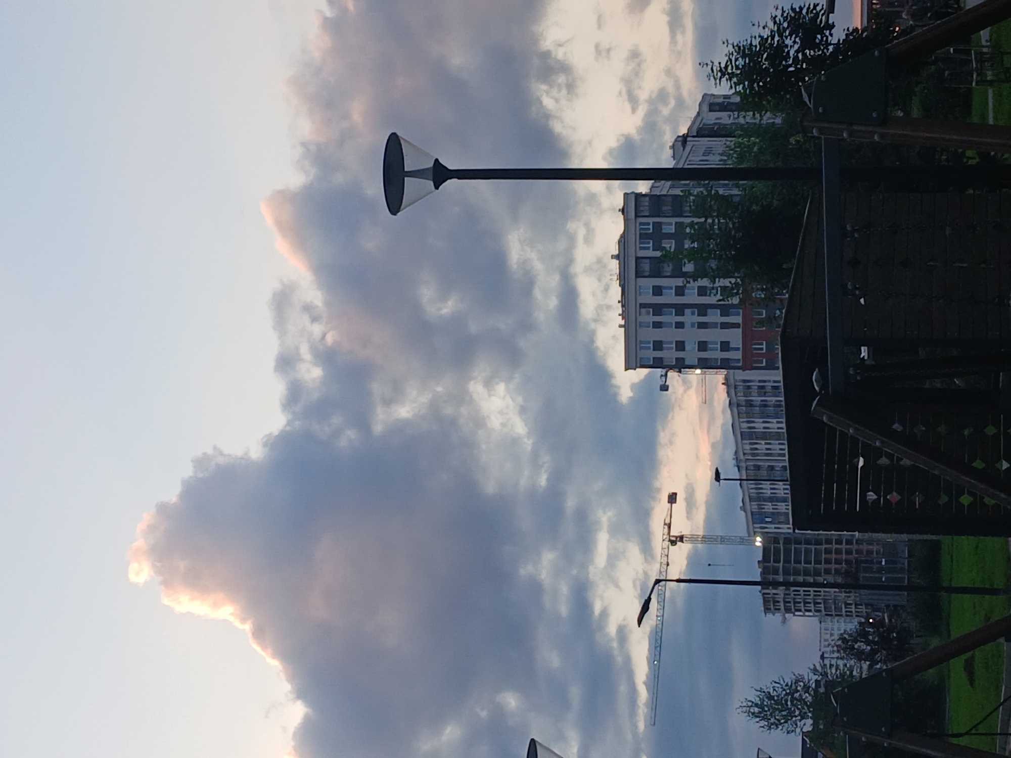 The image captures a dusk scene in Murino, Russia, featuring a mix of modern architecture and construction sites. In the foreground, a dark wooden playground structure with green accents is partially visible. A single streetlamp with a translucent shade stands prominently on the right side of the frame, its dark pole extending upwards. In the mid-ground, several residential buildings are under construction, some with visible scaffolding and concrete frames, while others are more complete with multi-story facades. A tall construction crane with a yellow boom is visible to the left, its arm extended. In the distance, completed residential buildings with numerous windows form a cityscape. The sky is filled with dramatic, heavy clouds, some illuminated with soft pink and orange hues, suggesting the last light of sunset. The overall impression is one of an urban environment in transition, with ongoing development set against a moody sky.