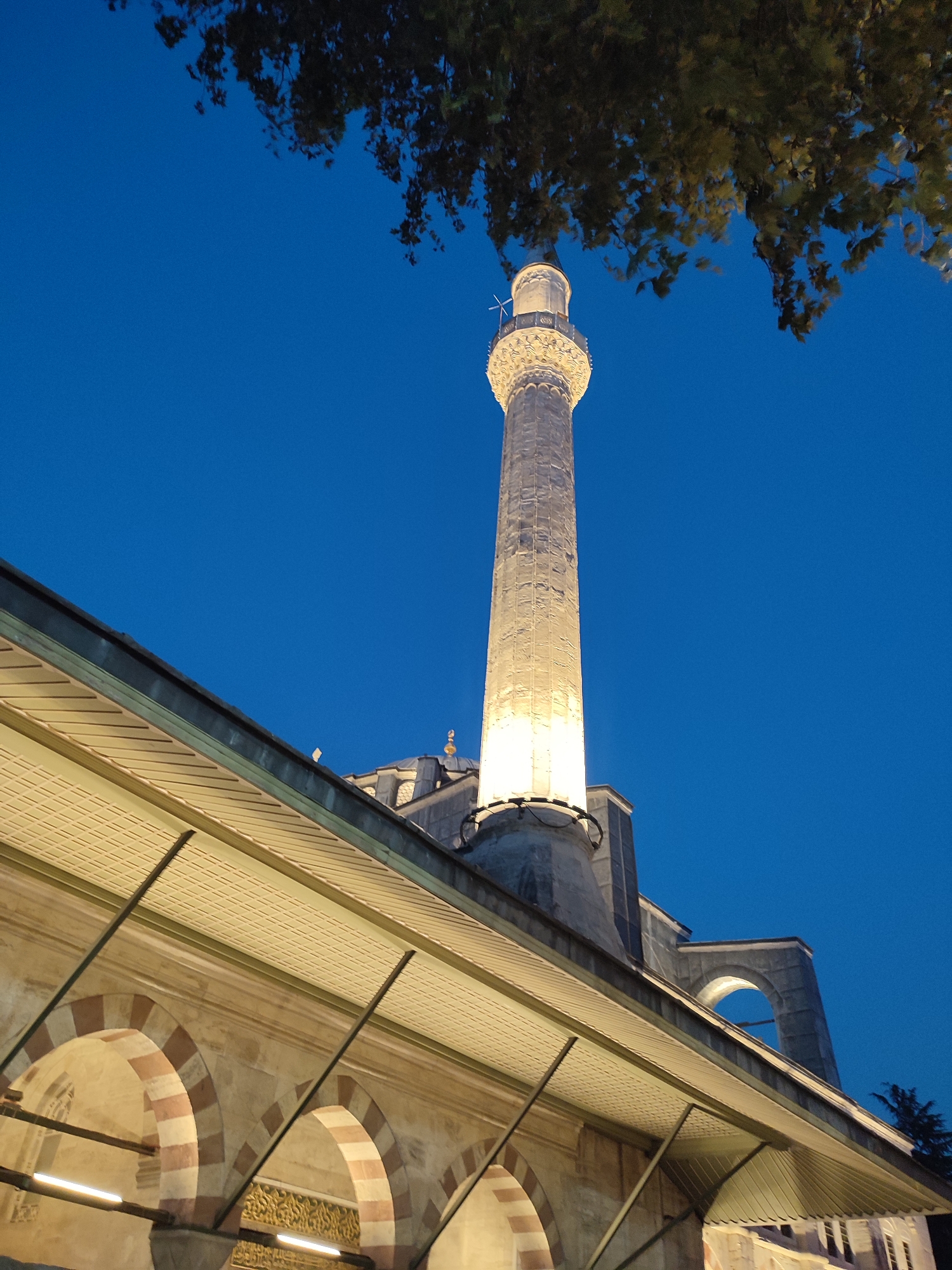 The image captures a minaret of a mosque at dusk in Beyoğlu, Turkey. The sky is a deep, clear blue, transitioning from twilight. The minaret, made of light-colored stone, is illuminated from below, casting a warm glow that highlights its detailed architectural features, including a balcony and decorative elements near its top. In the foreground, the roofline of a mosque building is visible, with a series of arched walkways supported by striped pillars. The ceiling of this walkway is made of a textured, off-white material, with angled beams providing support. Some tree branches, dark against the blue sky, hang into the frame from the top left. The overall scene conveys a sense of peacefulness and historical grandeur. There are no people visible in the image.