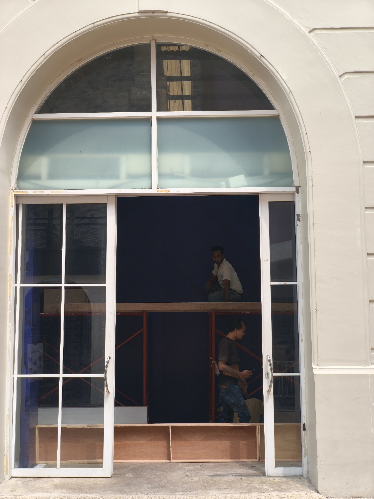 Two men are working on a construction project inside a large arched doorway in South Jakarta, Indonesia. The doorway has a white frame and features a large arched window above a set of glass double doors. The upper part of the doorway is filled with frosted glass panes, while the lower part consists of clear glass doors with a white grid.
One man, wearing a light-colored t-shirt and blue jeans, is crouched on a wooden platform inside the doorway, looking to his left. He appears to be supervising or inspecting the work. The second man, wearing a dark t-shirt and blue jeans, is standing further inside the doorway and to the right. He is holding a mobile phone in his right hand and looking down, possibly taking a break or communicating with someone.
Behind the men, scaffolding structures are visible, along with dark blue tarpaulin or fabric covering the back wall of the interior space. Wooden shelves or boxes are arranged on the floor within the doorway. The exterior of the building is a light beige color, with architectural molding around the arched doorway. The ground in front of the doorway is concrete.
