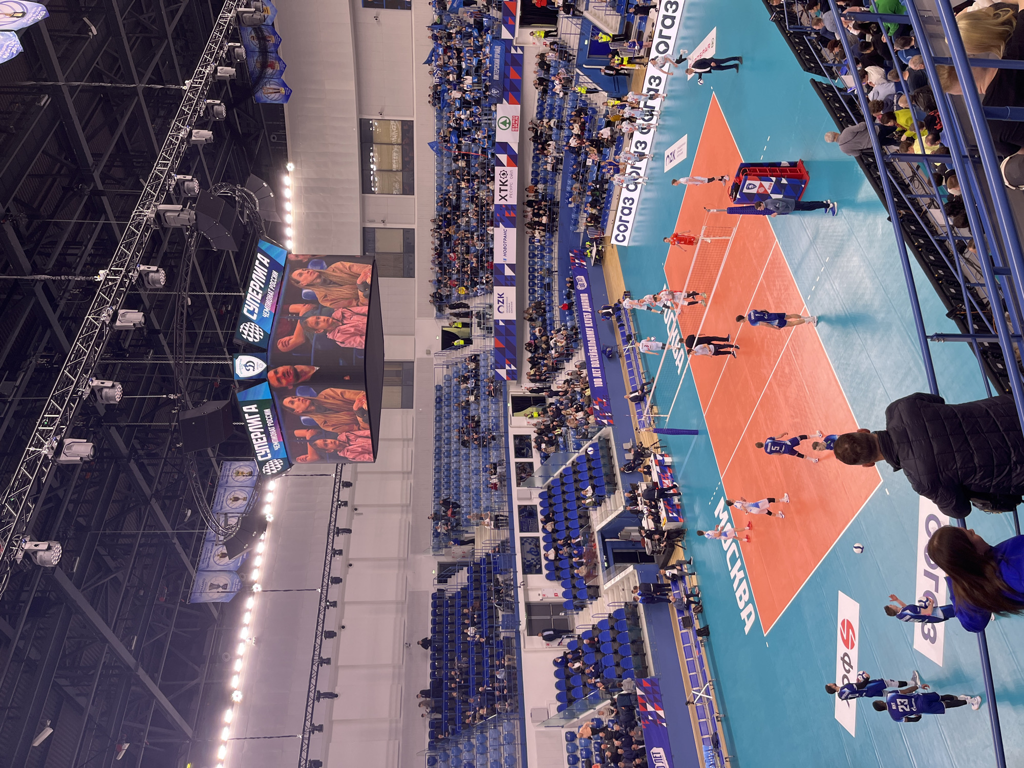 This image depicts a volleyball match taking place indoors in Moscow, Russia. The court is divided into two sides with a net in the center. Players in blue and red uniforms are actively engaged in the game. Spectators are seated in the stands around the court, and banners with corporate logos are displayed along the sides. A large screen above the court shows the match. The environment is a sports arena with lighting and rigging visible in the ceiling.