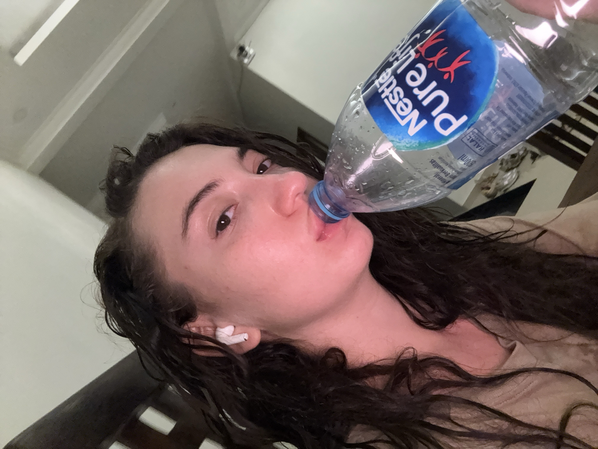 A woman with dark hair and fair skin is drinking from a "Nestle Pure Life" water bottle. She is wearing a white wireless earbud in her left ear. The picture appears to be taken at an angle, with a partial view of a room in the background, including a ceiling fixture and a wooden slatted structure. It is possible this image was taken in Dalung, Indonesia.