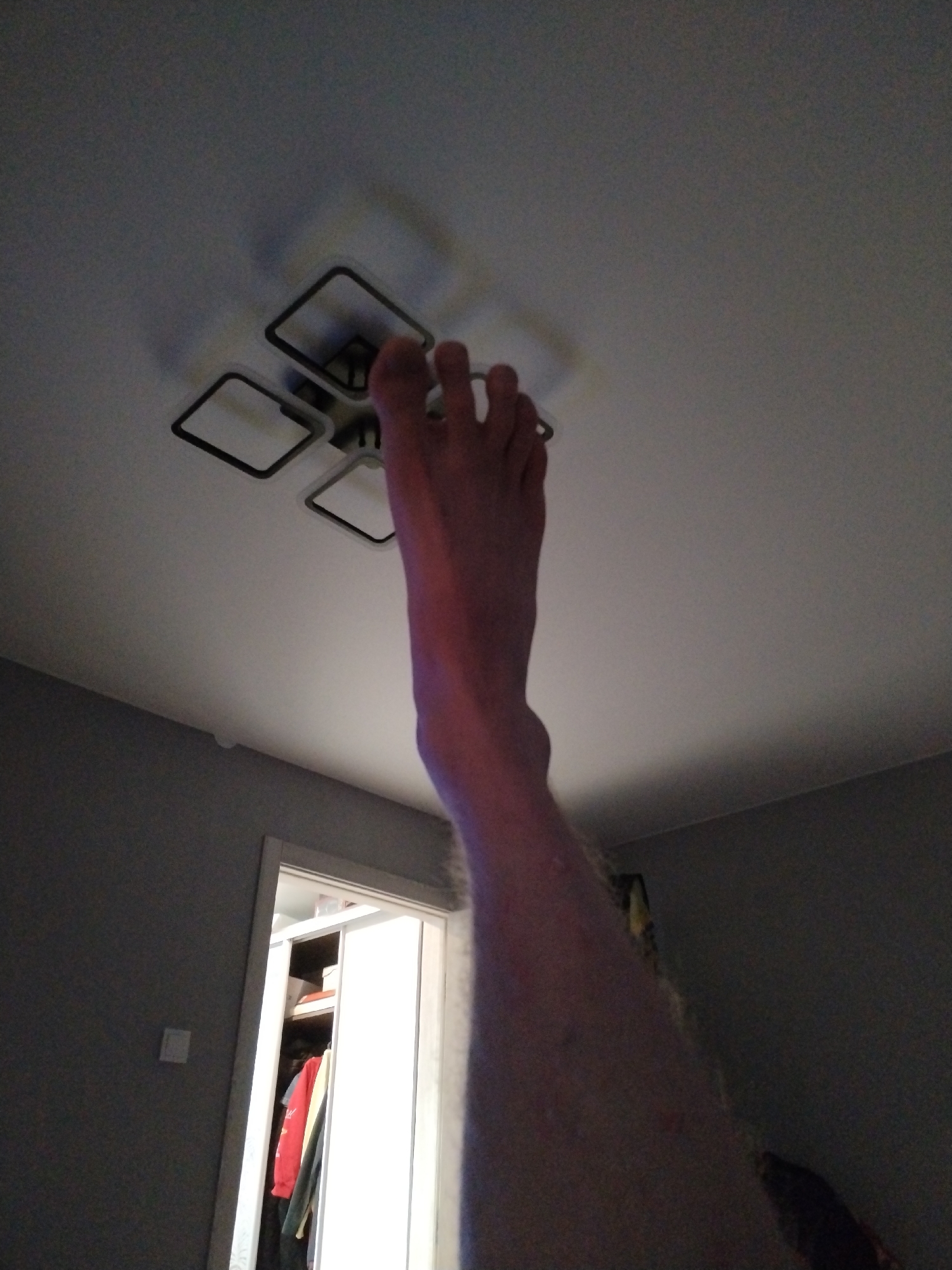 A photograph taken from a low angle shows a human foot reaching toward a ceiling light fixture. The foot's toes are close to the light. The leg extends downward into the frame. In the background, there's a partly open door revealing a closet or storage space with clothes visible inside. The walls are a dark gray color, and the ceiling is white. The location is not ascertainable from the image.