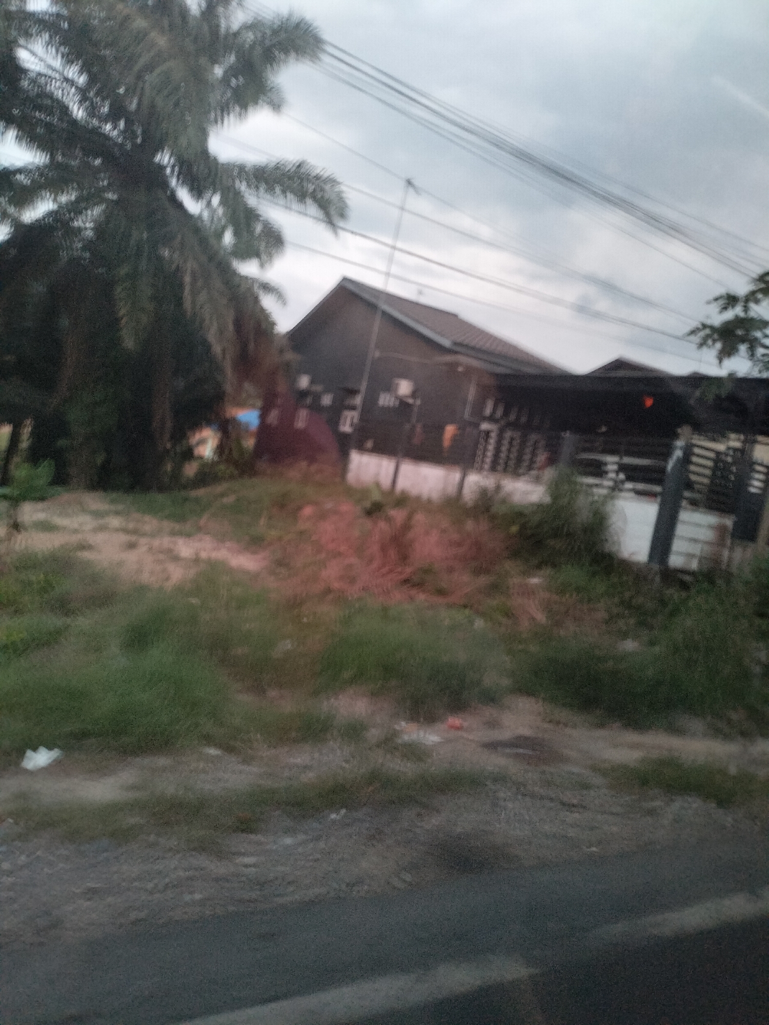 A blurred image shows a dark, wooden-sided building with a tiled roof in Bukit Selamat, Indonesia. A large palm tree is visible to the left of the building. The building appears to be a house or a shop. The area in front of the building is overgrown with grass and there are some scattered items on the ground, including what looks like a white plastic bag. Power lines crisscross the cloudy sky above. The scene suggests a rural or suburban setting.