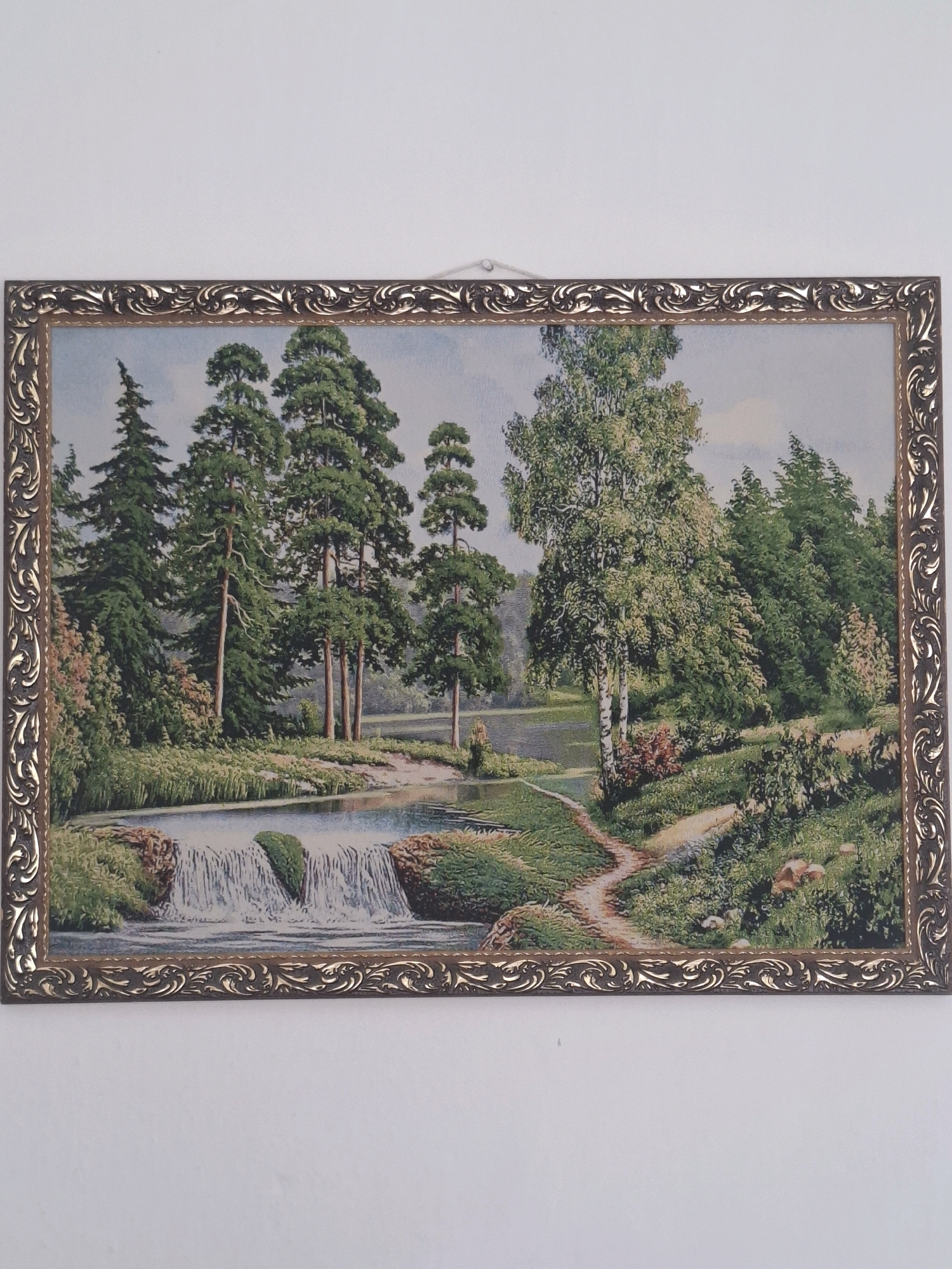 This is a framed tapestry depicting a landscape scene. The tapestry features a river with a small waterfall in the foreground, surrounded by lush green trees and vegetation. A pathway winds through the scene, and there appears to be a body of water in the background. The frame is ornate and decorative, with a metallic finish. The tapestry is hung on a plain white wall. There are no people visible in the image. The location in the city of Almaty, Kazakhstan, is not discernible from the image itself.