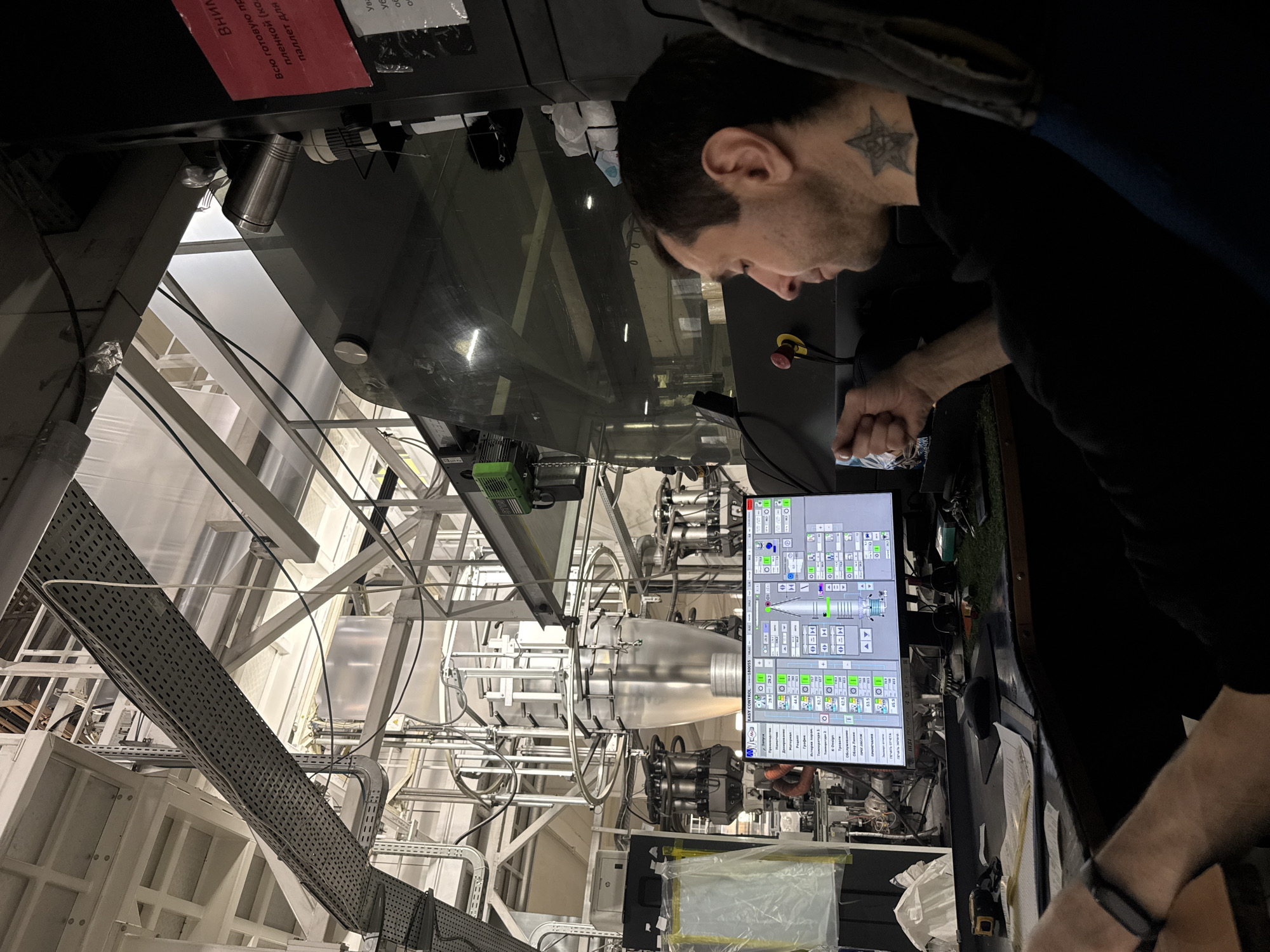 A man with a star tattoo behind his ear is operating a machine, likely in a factory setting, possibly for plastic film production. He is looking at a computer monitor with numerous control buttons and readouts. The machine behind the monitor features various tubes, wires, and a large, transparent, bubble-like structure. The location is unknown as there is no information about Perm, Russia.
