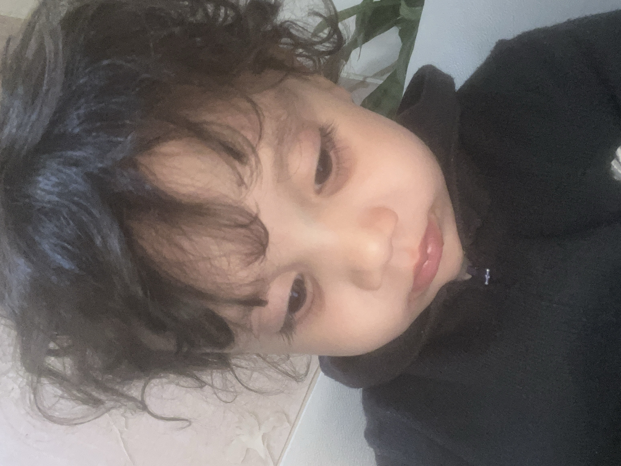 A close-up, angled shot captures a child with curly dark hair and long eyelashes. The child is wearing a black hooded sweatshirt, the zipper visible near their chin. Their skin tone appears light, and their eyes are directed downwards. A hint of green foliage is visible in the upper right corner, possibly from a plant. The background includes a white surface with a slight pattern. The location is Unknown, Unknown.