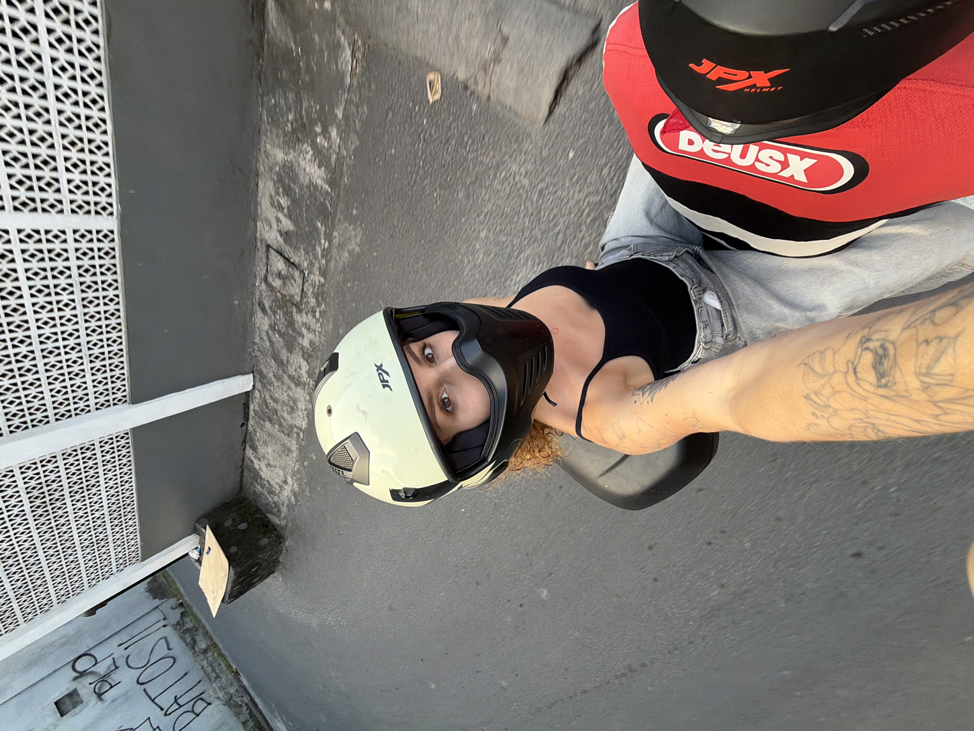 An eye-level, wide-angle selfie shows a woman wearing a light-colored motorcycle helmet with a black face shield and a black tank top, sitting on the back of a motorcycle. She has curly brown hair and is looking at the camera. Her left arm is extended towards the viewer, revealing tattoos. The motorcycle is moving on a wet asphalt road. To the right of the woman, a red and black object, likely a motorcycle fairing or luggage, is visible. In the background, a gray wall with white patterned ventilation blocks and graffiti on a white wall are present. A dark, cube-shaped object with a piece of wood on top is situated near the wall. The scene appears to be in an urban environment, possibly in North Kuta, Indonesia, given the architectural style and road conditions.