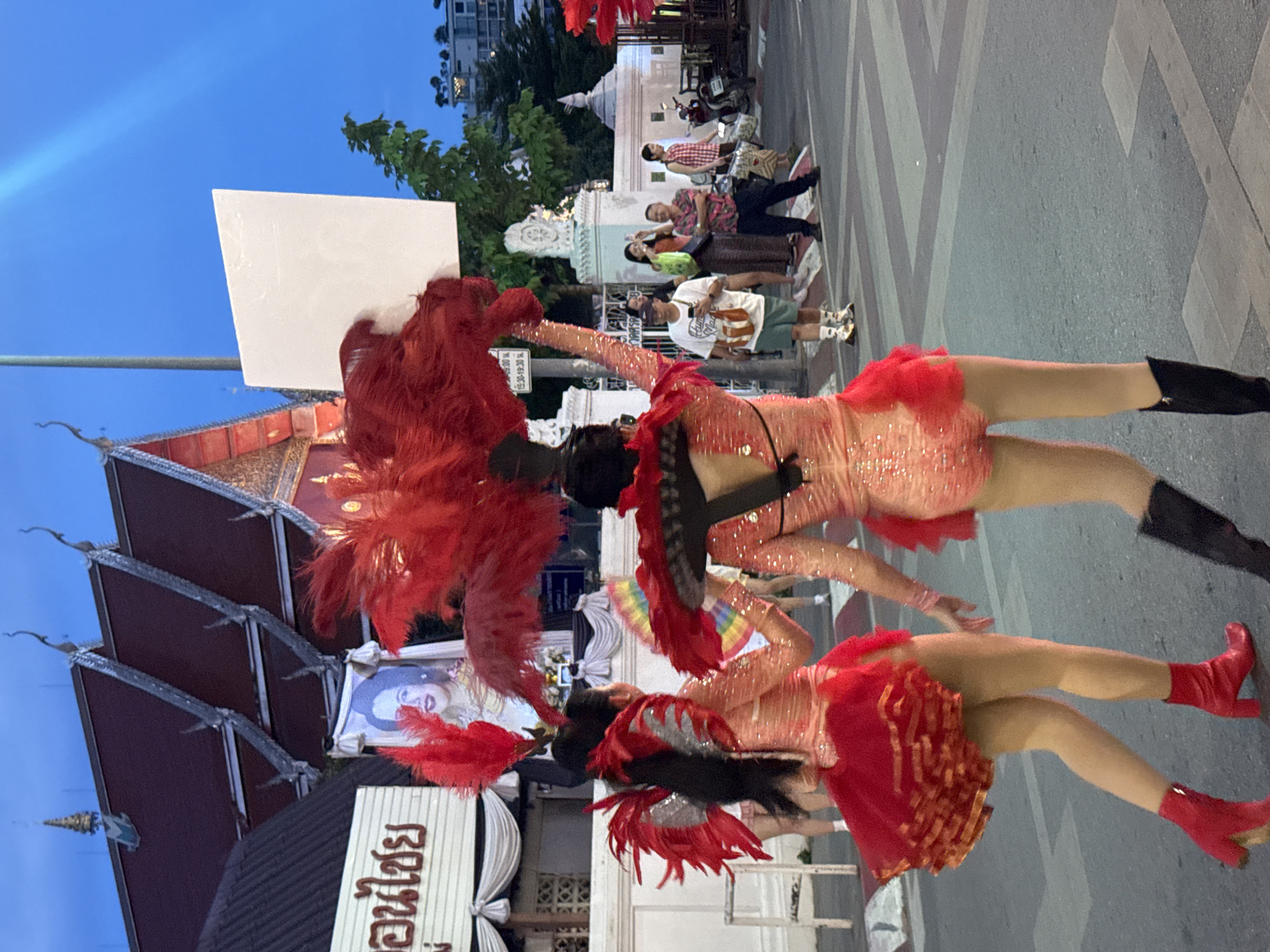 This image captures a vibrant street scene in Chiang Mai City Municipality, Thailand. Two individuals are prominently featured, seemingly performers dressed in dazzling, sequined pink dresses adorned with red feather accents. One wears tall black boots, while the other is in red ankle boots with heels. They appear to be posing or performing, possibly for a local event or celebration.
In the background, a group of people are observing or watching the performance. They are dressed in casual clothing and appear to be interacting amongst themselves. Behind them, there's a row of parked scooters.
On the left side of the image, there's a temple-like building. Next to it there is a sign in Thai and a portrait of a woman is hung. In the upper left corner, there's a white rectangular sign. The environment suggests an urban setting with a blend of cultural landmarks and modern elements. The lighting implies it may be dusk or evening.