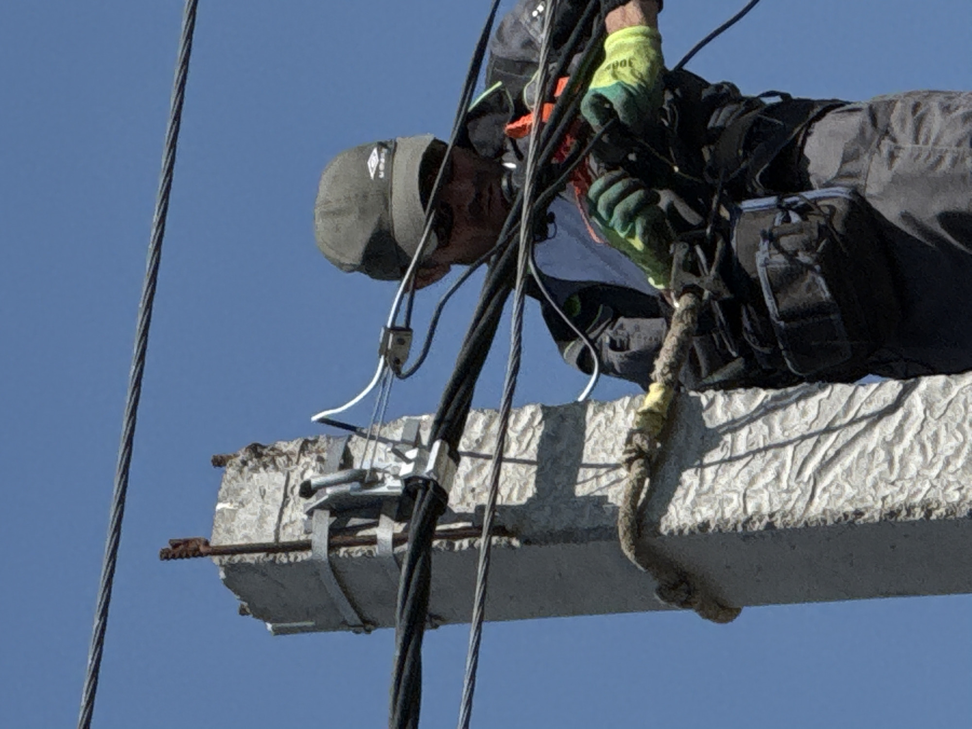 A man in a grey cap, work gloves, and a black jacket and pants is suspended in the air, working on a grey, weathered concrete beam. The beam is secured by metal straps and fittings. Black cables run vertically near the beam. The background is a clear, blue sky. The location is Unknown, Unknown.
