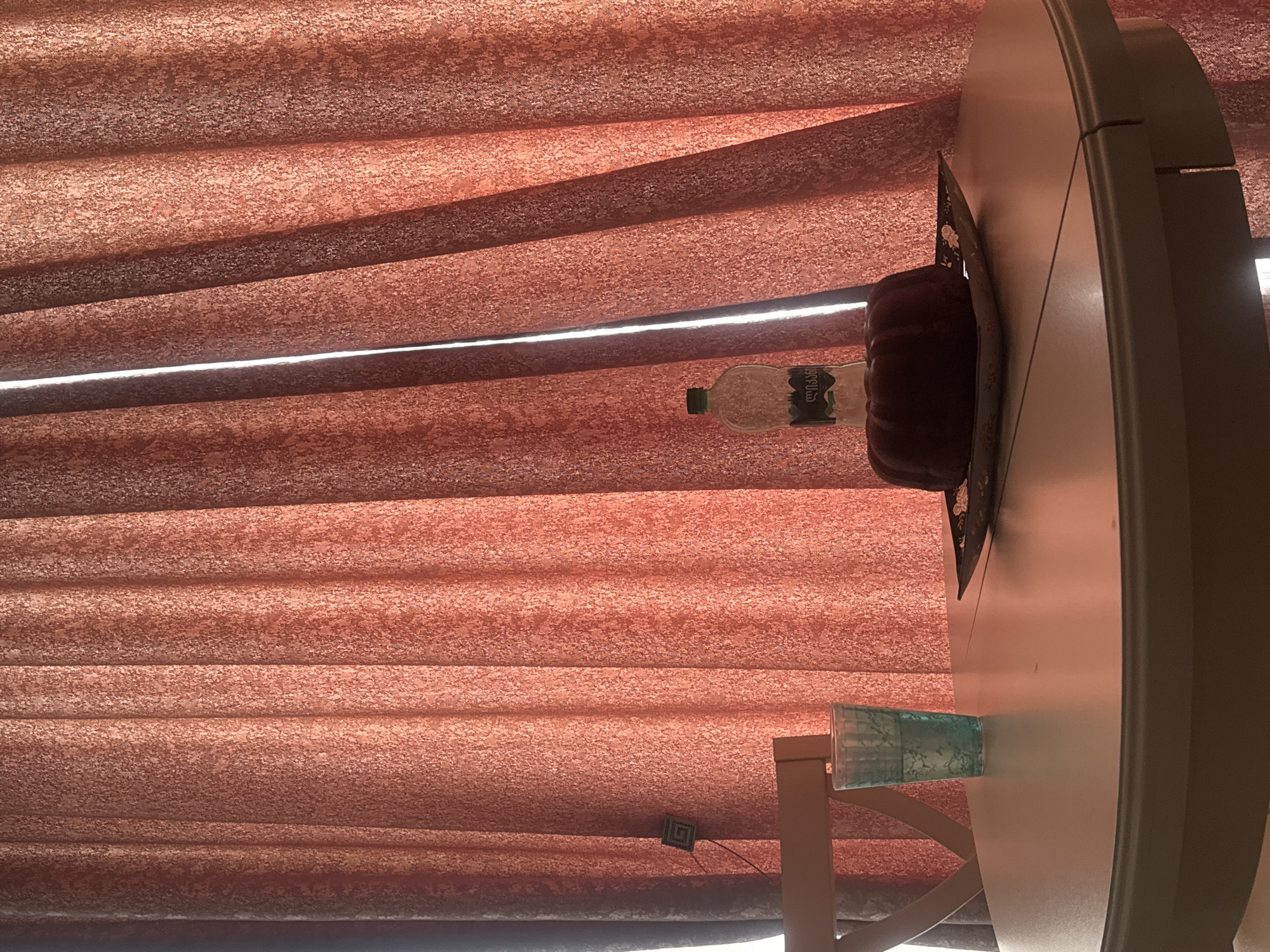 The image displays an indoor scene featuring a window with reddish-brown, textured blinds. The blinds are slightly parted, revealing a bright horizontal strip of sunlight. To the right, a light-colored, round table occupies a significant portion of the frame.
On the table, several objects are visible. A clear plastic bottle with a green cap contains a transparent liquid, and its label clearly displays the Georgian word "წყალი," meaning "water." Next to the bottle, a dark, fluted object, possibly a bundt cake, rests on a patterned mat or plate. Further along the table, a textured blue or teal glass is placed. Part of a wooden chair with a criss-cross back design is also visible on the far right. A small, square decorative item featuring a black and white spiral pattern is attached near the bottom left, possibly to the blinds or a nearby wall.
The bright light indicates that it is daytime. The scene appears quiet and domestic, with no human subjects or explicit activities taking place. The Georgian text on the water bottle aligns with the provided location context of Tbilisi, Georgia.