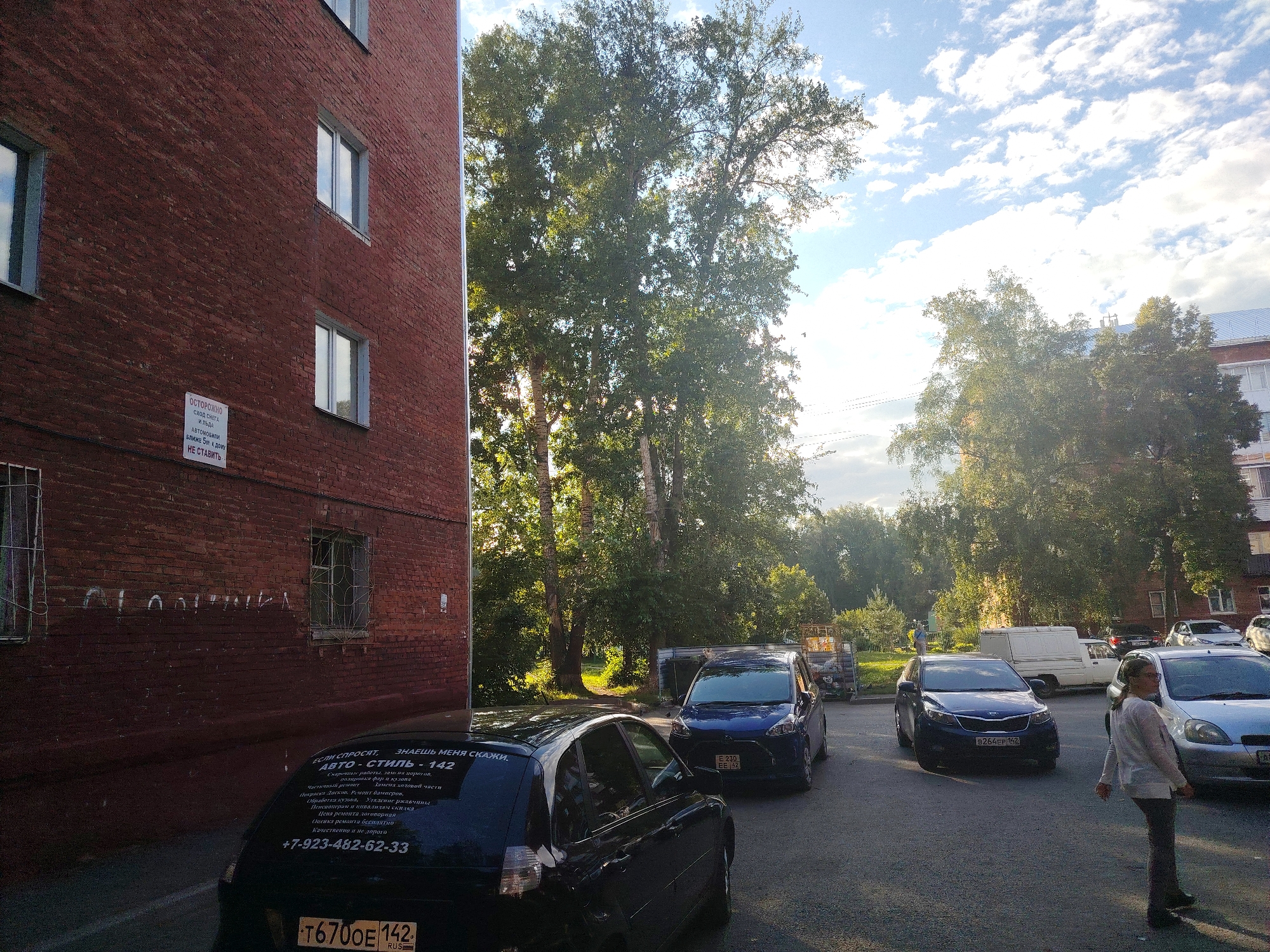 The image shows an exterior view of a residential brick building and a parking lot with several cars and trees in Kemerovo, Russia. On the left is a red brick building with several windows. A white sign with Russian text is affixed to the wall. In front of the building, several cars are parked. A black car with visible Russian license plate "T6700E 142 RUS" and text on its rear window is in the foreground. Other cars include a blue Kia with license plate "B264EP 142". A woman in a light-colored shirt and dark pants is standing in the parking lot. In the background, a white truck and other vehicles are visible. Trees with green foliage are scattered throughout the scene, with sunlight filtering through them. The sky is partly cloudy.