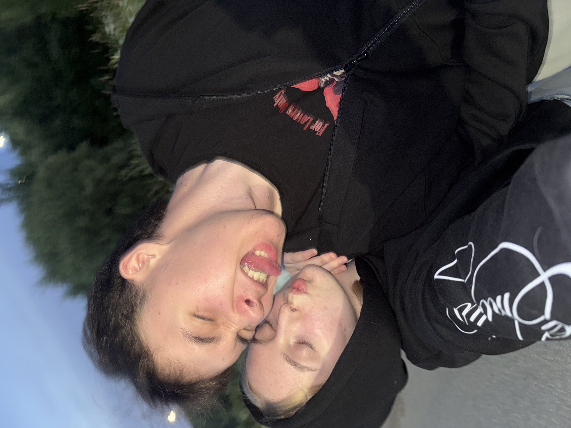 The image shows a close-up, portrait-style selfie of two individuals. The person in the foreground has their eyes closed and their tongue sticking out playfully. The second person is positioned behind and slightly to the side of the first, also with their eyes closed and their lips pursed, as if about to kiss. Both individuals are wearing black hoodies. The first person's hoodie has a red floral graphic and text that reads "For Lovers Only" printed on the chest, visible through the partially unzipped outer layer. The second person's hoodie has a white graphic design on the sleeve. The setting appears to be outdoors, with blurred greenery in the background and a pale sky. The lighting suggests it could be late afternoon or early evening. The overall mood is intimate and playful. The location context provided is Ryabinino, Russia.