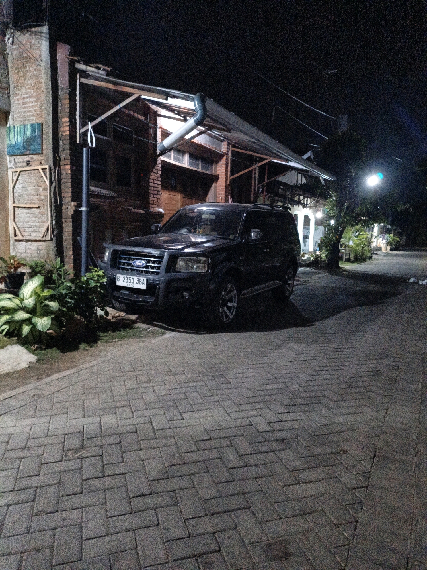 The image shows a black Ford SUV parked on a brick-paved street at night. The SUV has a license plate that reads "B 2351 JBA". To the left of the SUV is a brick building with a covered porch. There are plants in pots near the building. In the background, there are trees and a street light. The scene appears to be residential. The image does not provide specific details about the city of Cikal, Indonesia, beyond its implied presence in the license plate.