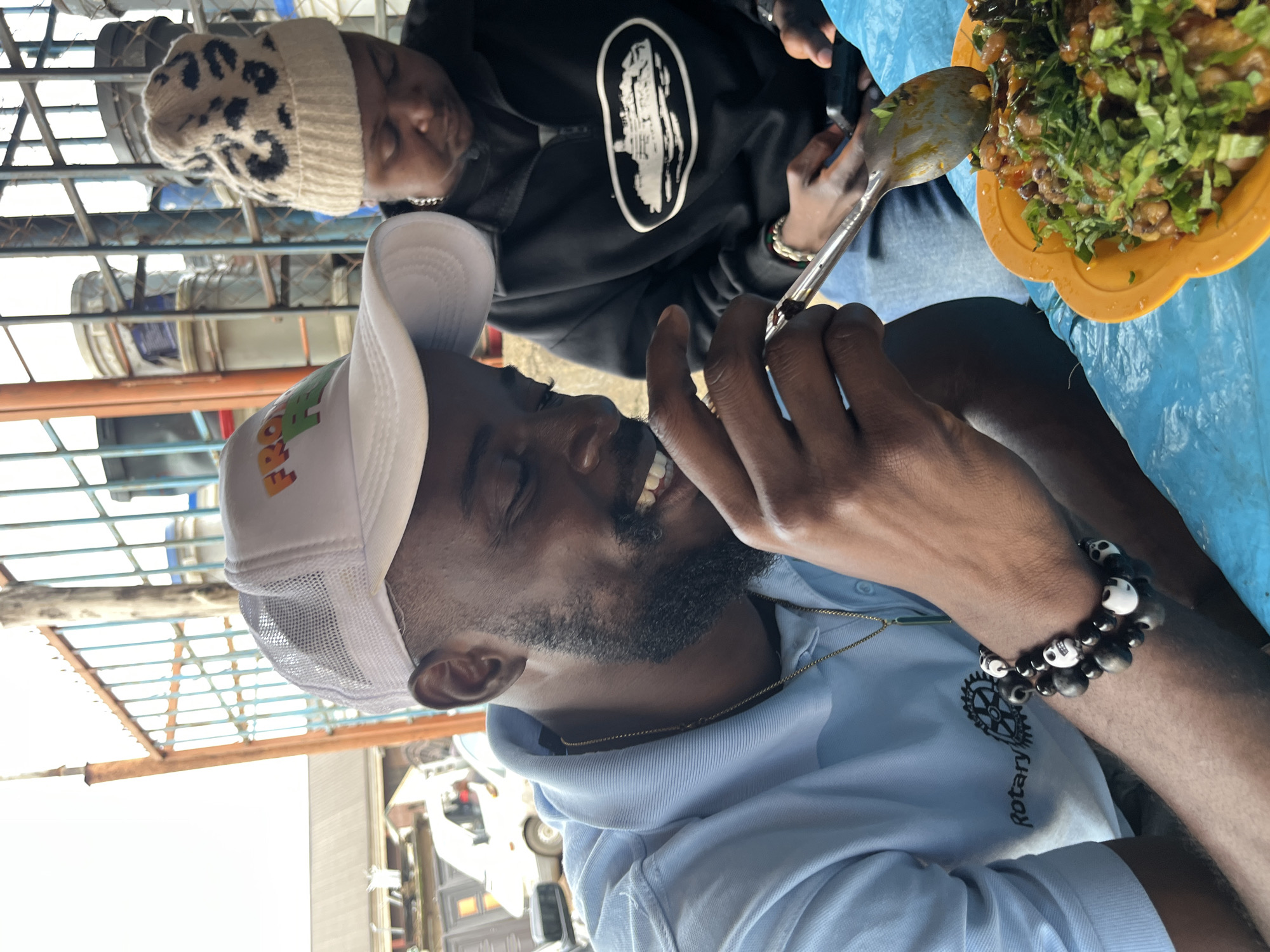 The image shows two people in an outdoor setting, possibly in Gura Top, Nigeria. The foreground features a man wearing a light blue polo shirt and a white cap with a mesh back. He has a beard and is smiling, looking at something in his hand. A gold chain is visible around his neck, and he is wearing two bracelets on his wrist, one with black and white skull-shaped beads. He appears to be holding a spoon, about to eat from a bowl of food. The food in the bowl is a mixture of beans and chopped green herbs, served in an orange plastic bowl.
Behind him, another person is visible, wearing a black hoodie with a white graphic design and a beige beanie with a leopard print pattern. This person is looking down at their hands, which are holding a phone and a metal spoon. The setting appears to be somewhat rustic, with metal bars and structures visible in the background, suggesting an open-air environment. The lighting suggests it is daytime. The overall mood is casual and relaxed, with the man in the foreground displaying an expression of contentment. The text "Rotary" is visible on the man's polo shirt.