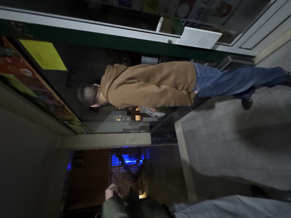 The image captures a nighttime outdoor scene in Bragança, Portugal, on a paved surface outside a commercial establishment.
Two individuals are present. On the right, a person is seen from behind, facing a shop window. This individual wears a brown hooded sweatshirt and blue jeans, with their right hand inserted into a pocket. Their left hand holds a white object or package near the lower edge of the shop window.
The storefront features a large glass display window and an adjacent glass-paneled door with a white frame. Above the display window, a colorful banner depicts various products, and a yellow rectangular sign is affixed to the upper right corner of the window. The address "Nº 15" is visible above the window. A white rectangular sign, displaying "MB" and "Bragança" within a red border, is attached to the glass door on the right.
To the left, a second person is partially visible, showing their upper torso, left arm, and left leg. This individual wears a dark olive-green jacket or hoodie and light grey pants. Their left hand is raised in front of their chest.
In the background, behind the left individual, an outdoor staircase leads to a building. A prominent blue light source illuminates the area behind and beneath this staircase, creating reflections on the ground. The ground is a light grey tiled surface, exhibiting shadows cast by the figures and ambient structures.