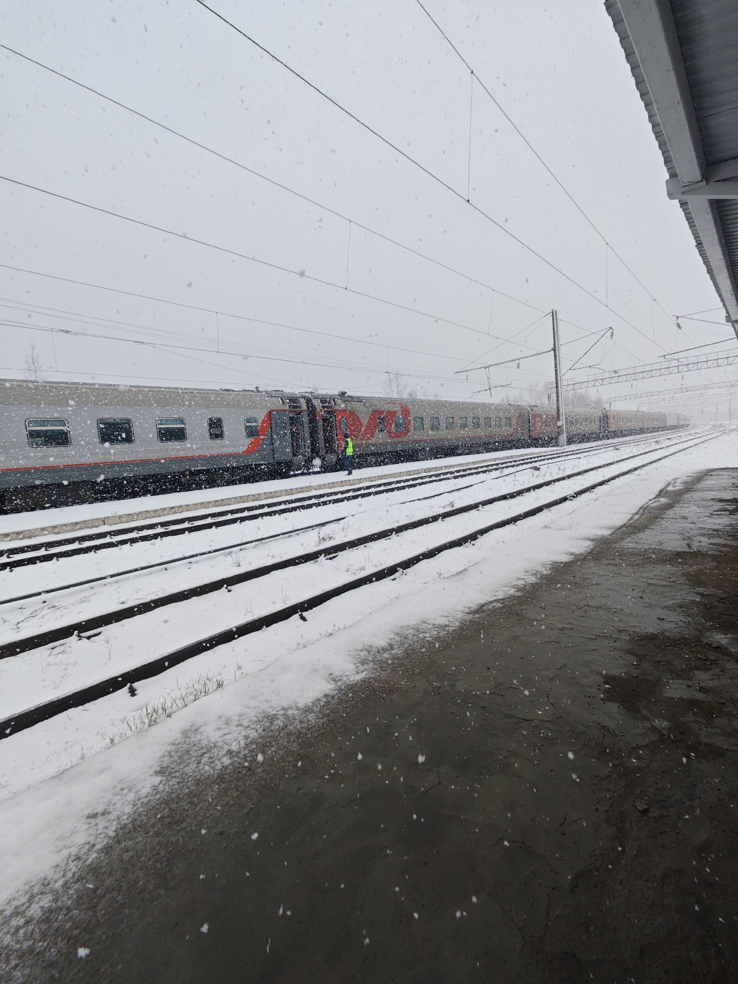 The image depicts a train at a station in Kambarka, Russia, during a snowy day. The train cars are silver with red accents and the Russian Railways logo is visible. A person in a yellow vest stands near an open train car door. Snow covers the train tracks and the ground adjacent to the platform, which is partially wet. Overhead power lines run above the train. The sky is overcast with snow falling.