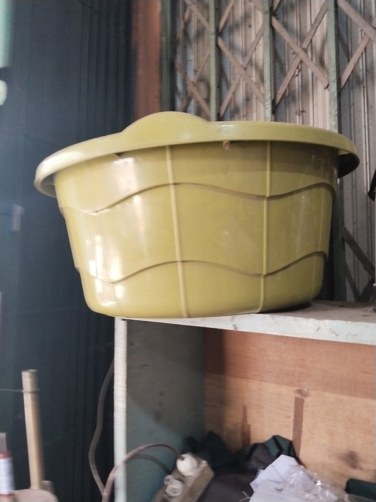 A pale yellow plastic basin rests on a shelf in what appears to be a storage area or workshop. The basin has a ribbed design around its exterior. Behind the basin, a grey metal accordion-style gate is visible, and to the left, a dark blue wall with vertical lines. The shelf itself is made of a light-colored material, possibly concrete, with a wooden surface underneath. The overall environment suggests a functional, possibly industrial or storage setting.