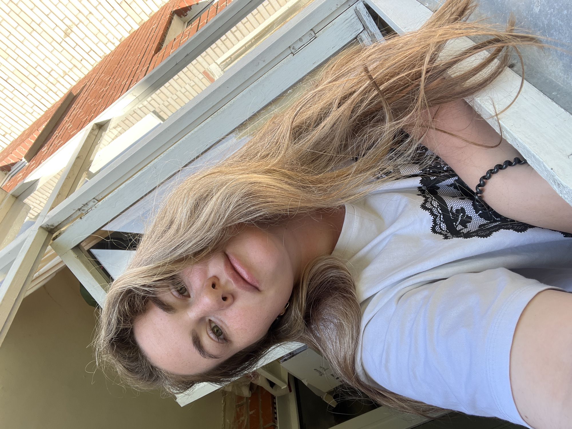 A young woman with long, blonde hair is positioned in front of a window frame. She wears a white T-shirt with black lace detailing on the shoulder area and a black spiral hair tie is around her wrist. The window frame is painted white, and a brick building is visible through the window. The exact location is implied to be Chaikovsky, Russia.