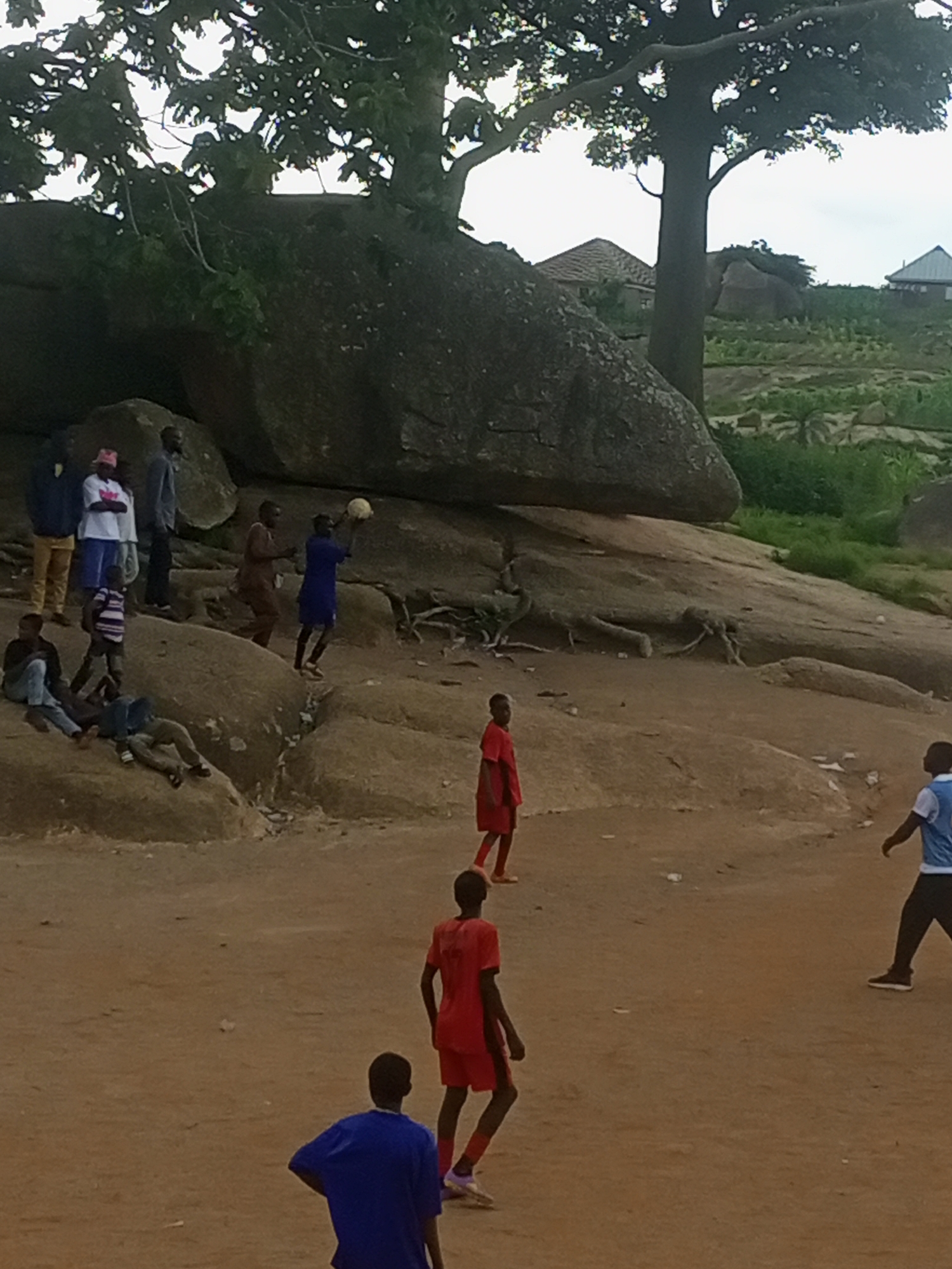 In the city of Jos, Nigeria, a group of young men and boys are gathered on a rocky terrain near some trees. The scene appears to be an outdoor recreational area. In the foreground, a young person in a blue shirt is facing away from the camera, while another in a red shirt is looking to the side. Further back, a group of individuals are interacting. One person, dressed in a blue shirt, is holding a ball above their head, suggesting a game is in progress or about to begin. Several other people are standing or sitting on the rocks. The environment is characterized by large rocks, dirt ground, and sparse vegetation. In the background, there are structures that appear to be houses with thatched roofs, indicating a semi-rural or residential area on a hillside. The sky is overcast.