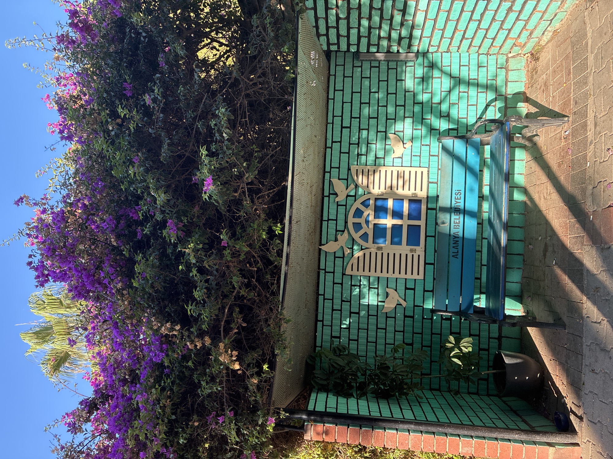 The image depicts an outdoor seating area under a canopy of vibrant purple bougainvillea flowers and a palm tree. The seating area is constructed with turquoise brick walls and a patterned brick floor. A blue metal bench with "ALANYA BELEDİYESİ" inscribed on it is placed against the back wall. Above the bench, a decorative window with shutters and blue panes is mounted, flanked by three white bird figures. A potted plant sits to the left of the bench. The scene is illuminated by sunlight, casting shadows on the ground.