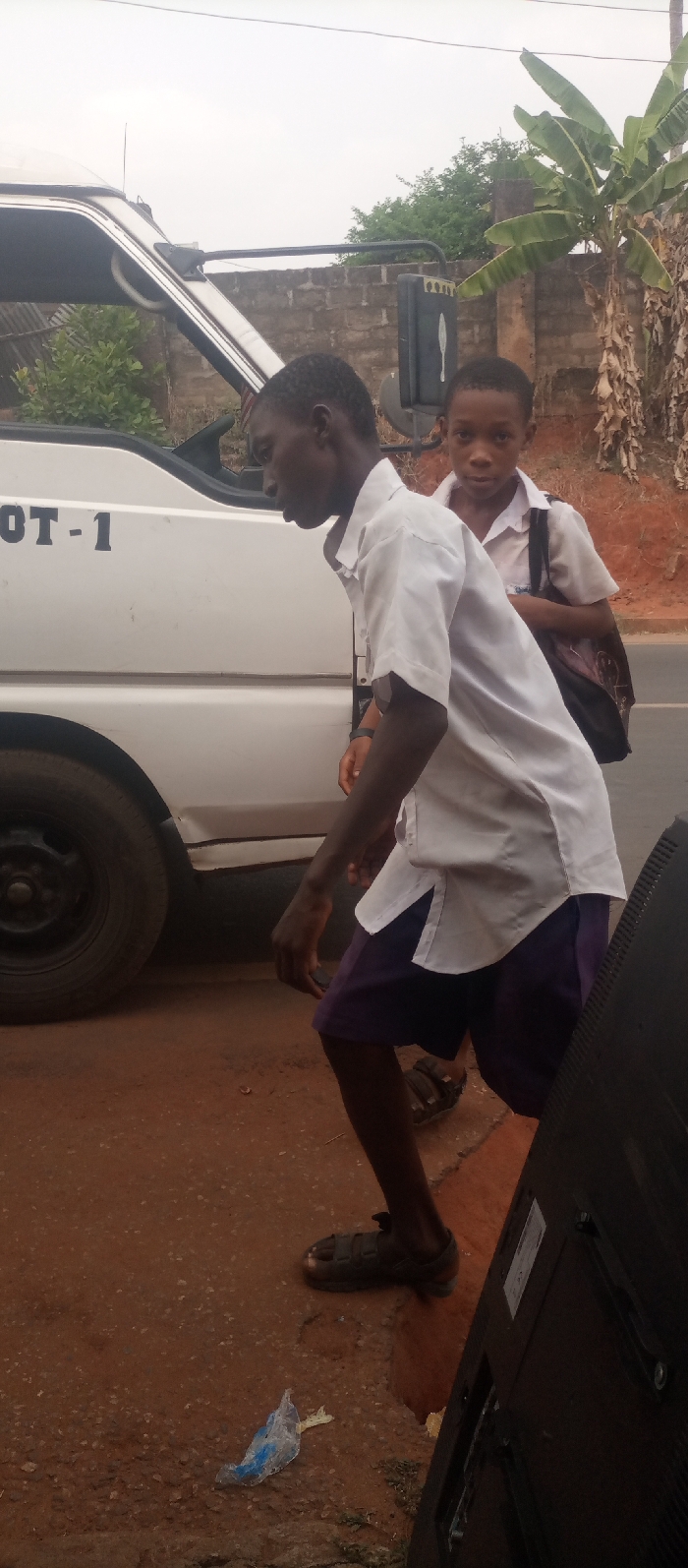 The scene depicts an outdoor area in Ogidi, Nigeria, possibly a roadside. Two dark-skinned males are present. In the mid-ground, an adolescent male wears a white short-sleeved collared shirt, dark purple shorts, and dark sandals. He is positioned left of center, leaning slightly forward and facing left, with his right arm bent. Behind him and to the right stands a younger male child, wearing a white short-sleeved collared shirt and a dark backpack over his left shoulder; he looks directly at the viewer. A white van or mini-bus with the black letters "OT-1" on its side is partially visible on the left, showing its front tire and side panel. The ground consists of reddish-brown compacted earth. A crumpled light blue plastic wrapper lies on the ground near the adolescent male's feet. In the right foreground, a dark, rectangular object consistent with the rear of a television monitor stands vertically. The background features a low, light-colored cinder block wall, behind which green foliage and a large banana tree are visible. The sky is overcast and light grey.