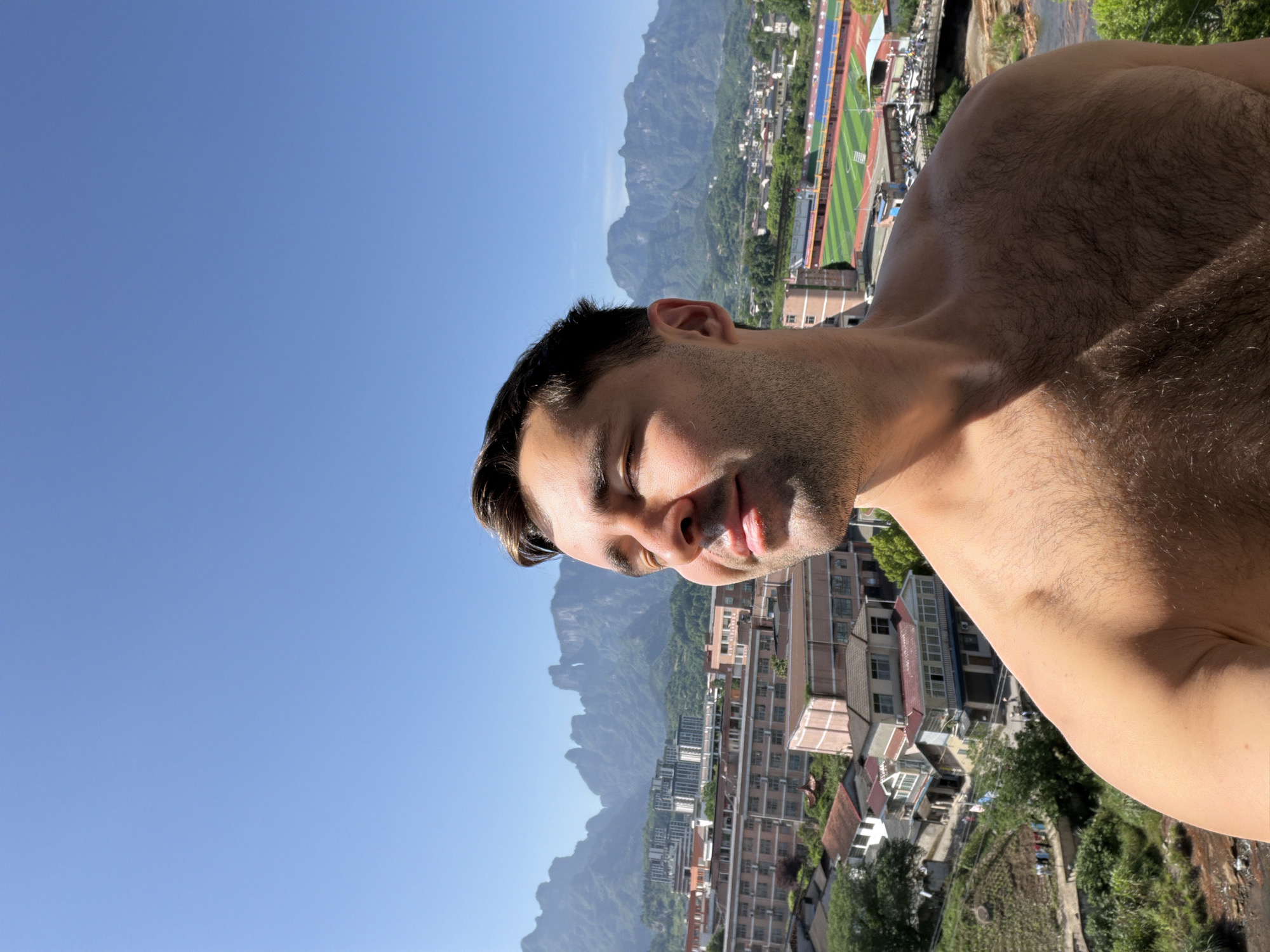A shirtless man with dark hair and a tan complexion is pictured from a low angle. His head is tilted down and to the side. His eyes are closed, and his tongue is slightly sticking out of his mouth. The background consists of a clear blue sky, mountains, and a city. The city has tall buildings, a running track with a green field, and other structures. The city appears to be Zhangjiajie, China, based on the unique mountain formations.