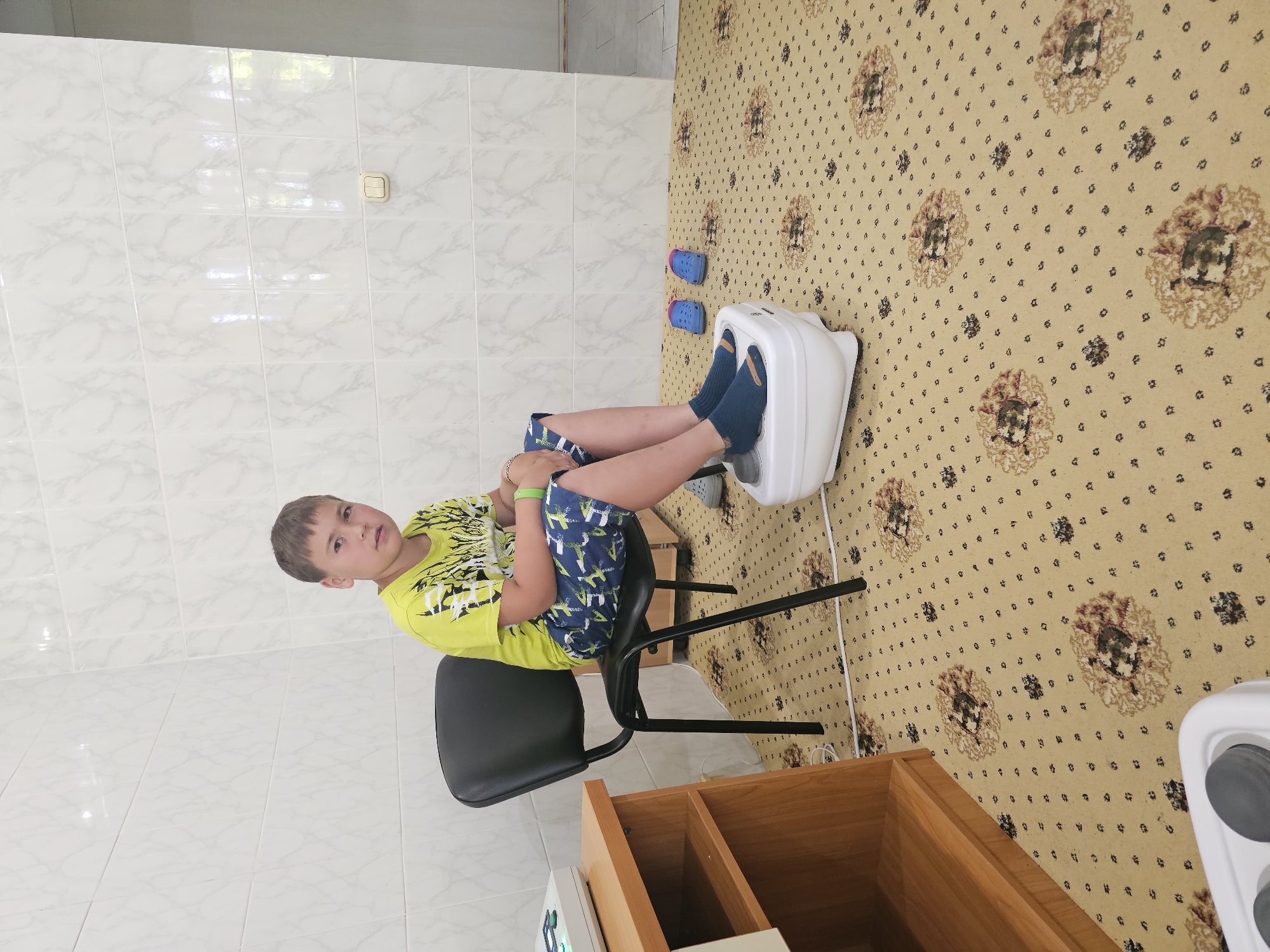 A young boy sits on a black chair in a room with tiled walls and a patterned carpet. He is wearing a bright yellow t-shirt with a graphic design and blue shorts with a white pattern. His feet are placed on a white foot massager. Blue slip-on shoes are positioned on the carpet next to the massager. To the left of the boy, a wooden shelf unit holds a medical device. The room appears to be a clinic or spa, possibly located in Lermontovo, Russia, given the visual cues which are consistent with common interior designs found in such facilities in the region.