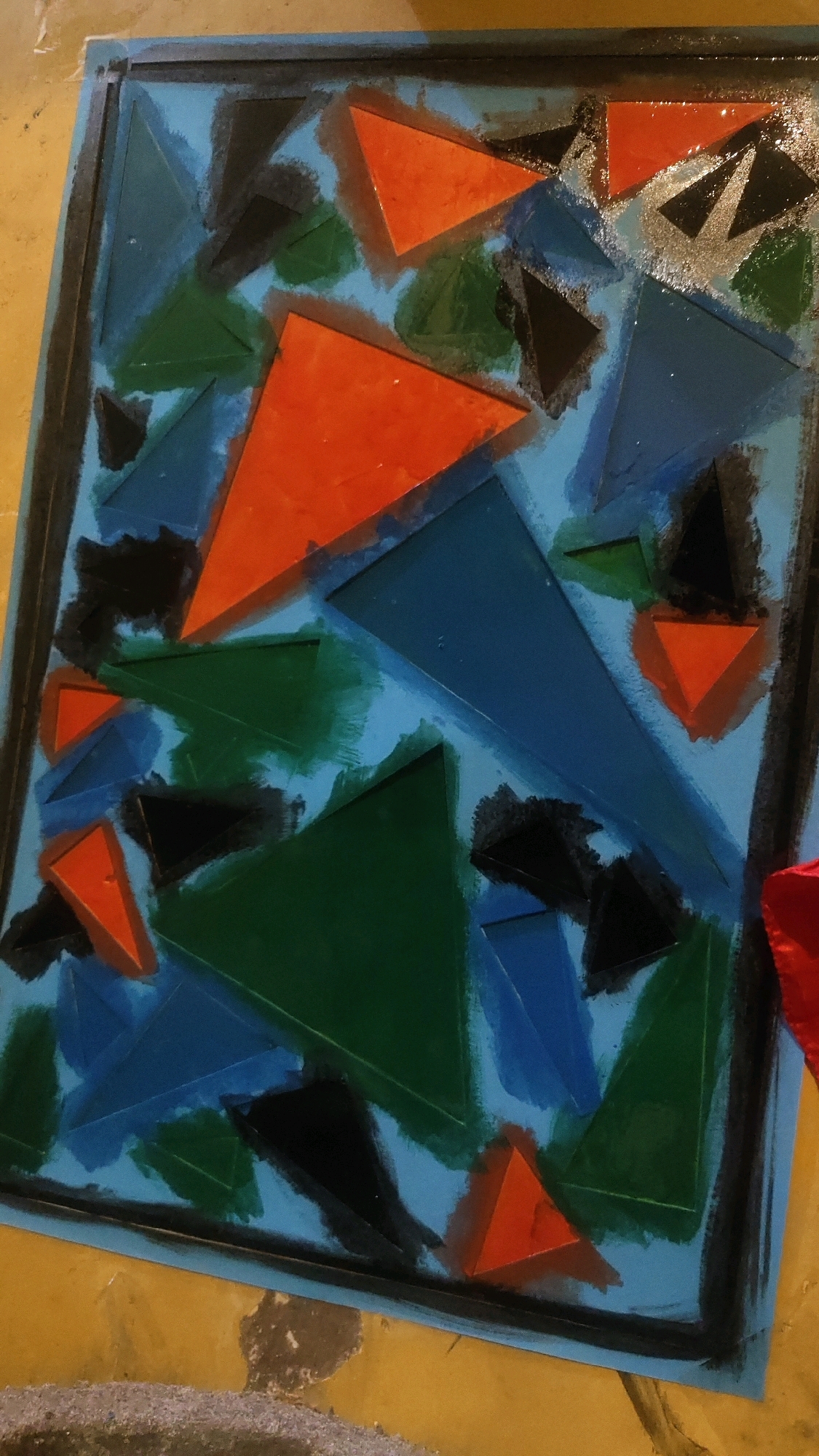 The image displays an abstract painting on a light blue canvas. The painting is characterized by a geometric composition of overlapping triangles in various colors including orange, green, blue, and black. Some triangles appear to be raised from the surface, creating a textured effect. The edges of the canvas are outlined with a thick black border, and the canvas itself rests on a yellow surface that appears to be textured and possibly worn. There are no people or specific landmarks visible that would indicate a location within Kediri City, Indonesia.