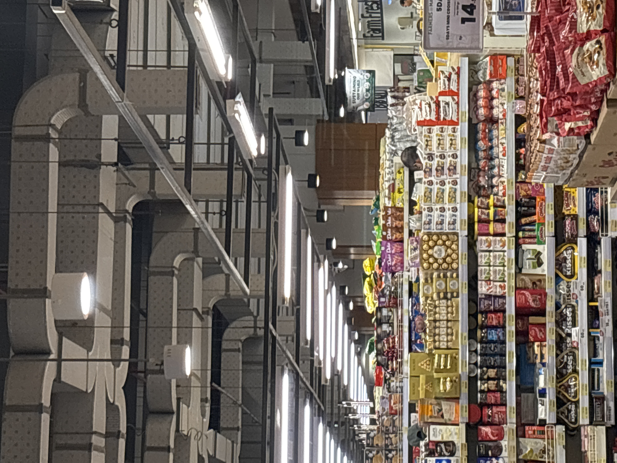 The image depicts the interior of a supermarket, possibly located in North Jakarta, Indonesia, based on OCR text from "Farm Fresh". The view appears to be from the end of an aisle, looking down its length. Rows of shelves are filled with assorted food products, including packaged snacks, chocolates, and baked goods. Shoppers are present along the aisle. Above the shelves, the supermarket's infrastructure is visible, including ductwork, lighting fixtures, and overhead signage.