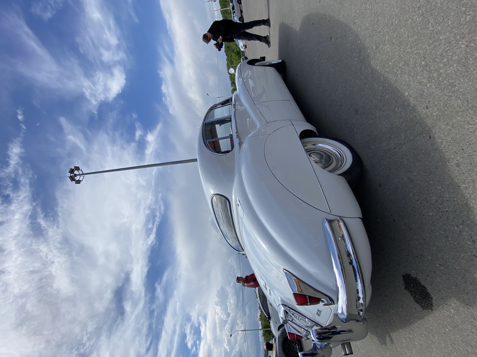 A white vintage Jaguar E-Type is parked outdoors on an asphalt surface under a cloudy sky. The car is viewed from the rear passenger side, highlighting its curved body, chrome bumper, and whitewall tires. A person in a dark jacket and pants is standing to the right of the car, holding a camera and appearing to take a photograph of the vehicle. In the background, another person in a red jacket is visible near a different car. A tall light pole with multiple lamps stands prominently in the sky. The scene suggests an outdoor car show or gathering.
