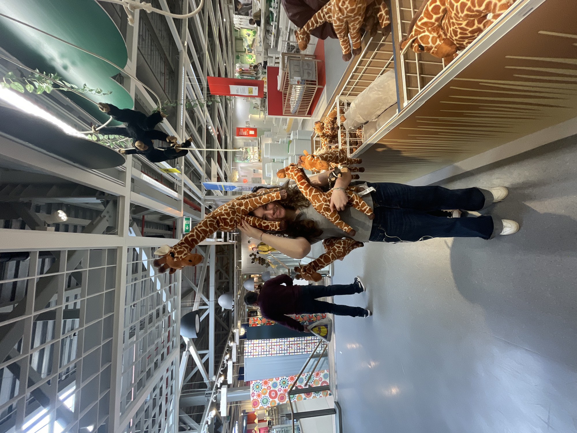The image shows the interior of an IKEA store, likely in Sofia, Bulgaria, given the common design and layout of IKEA stores worldwide. A young woman stands in the aisle, surrounded by several plush giraffe toys, some held around her head and neck. To her left, a man in a maroon sweater walks away. Overhead, more giraffe plush toys are displayed on shelving. The aisle leads to displays of furniture, bedding, and other home goods. The store's interior features a high, exposed ceiling with visible structural support. A hanging display of plush monkeys and artificial vines is also present. The floor is light gray, and the lighting is bright, typical of retail environments.