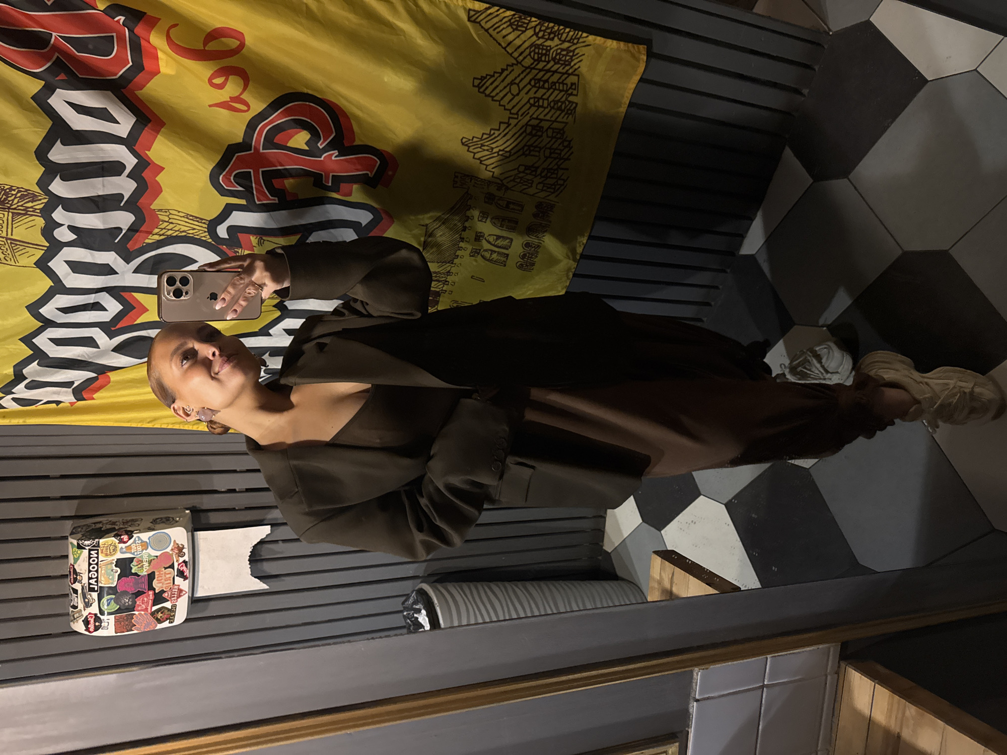 This image captures a mirror selfie taken indoors, likely in a public space such as a bar or cafe in Chaikovsky, Russia. The main subject is a person, appearing to be female, dressed in a dark olive-green long-sleeved top or blazer over a lighter brown undergarment and matching pants. She has short, light-colored hair pulled back and is holding a gold-colored iPhone in her right hand, positioned to take the picture. She has a slight, serene smile.
Behind the person, a large yellow banner or flag with bold red and black text and graphics hangs on a dark slatted wall. The text visible on the banner includes "Bomber" in a stylized, distressed font, and what appears to be "196" or "96L". Other smaller graphic elements resembling buildings or cityscapes are also present on the banner.
To the person's left, a white tissue dispenser covered in numerous colorful stickers is mounted on the slatted wall. Visible text on the stickers includes "LAGOON", "GHOST", "BLISS", and "JUICY CRUSH". Below the banner and behind the person, the floor features a striking geometric pattern of black, white, and grey hexagonal or octagonal tiles. The person is wearing light-colored, chunky sneakers. The overall lighting suggests an indoor, artificial light setting, possibly in the evening.