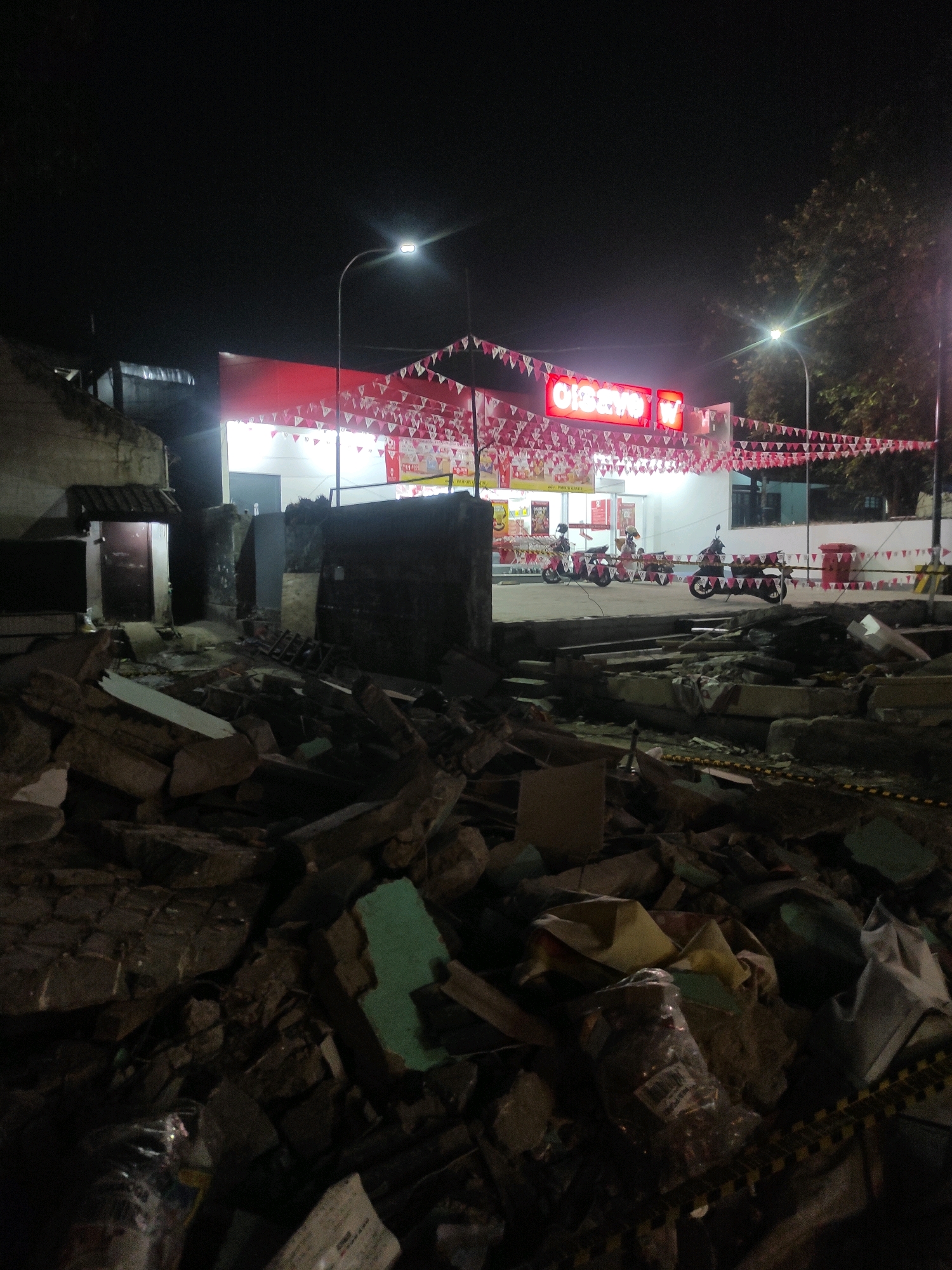 The image shows a nighttime scene in Padalarang, Indonesia, depicting a commercial establishment with a red facade and neon signage that reads "CISSOYO". The entrance is decorated with red and white triangular pennants. Several motorcycles are parked in front of the building. In the foreground, there is a significant amount of rubble and debris, suggesting a recent demolition or destruction. The rubble consists of concrete, bricks, and other construction materials, with some plastic sheeting and bags visible among the debris. The scene is illuminated by streetlights and the lights from the commercial building, casting long shadows. The overall impression is one of contrasting destruction and ongoing commerce.