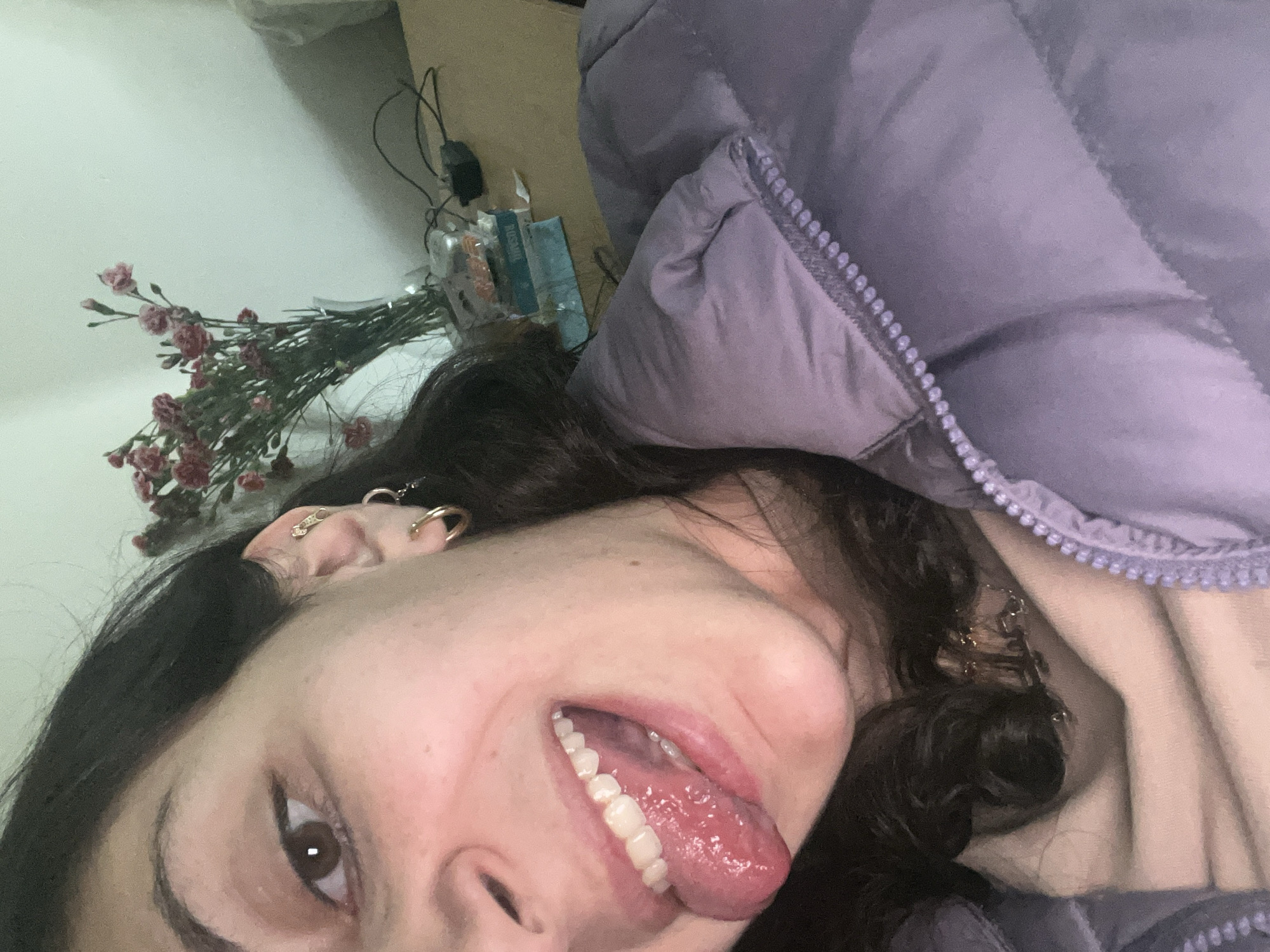 This image captures a close-up, high-angle selfie of an individual taken indoors, located in Mexico City, Mexico. The subject's face is partially visible, encompassing the right side from the nose down, their left eye, and extending to the upper chest. They possess dark brown eyes, a defined left eyebrow, and dark, curly hair. The visible left ear features multiple piercings, including a gold-colored hoop in the lobe, a smaller gold-colored hoop or stud above it, and a distinctive gold-colored zipper-shaped earring in the helix. The individual's mouth is open, with the tongue extended forward, appearing moist and pink. White teeth are visible. The subject wears a light beige or cream-colored garment beneath a quilted, light purple puffer jacket with a visible zipper partially open. In the background, a white wall is present, behind which sits a light-colored wooden surface, likely a table. On this surface, a clear glass vase holds a bouquet of small, reddish-pink flowers, resembling carnations. Adjacent to the vase, an electrical cord with a black adapter and several indistinct objects, including a blue box with white text, are visible. The scene is illuminated by soft, ambient light.