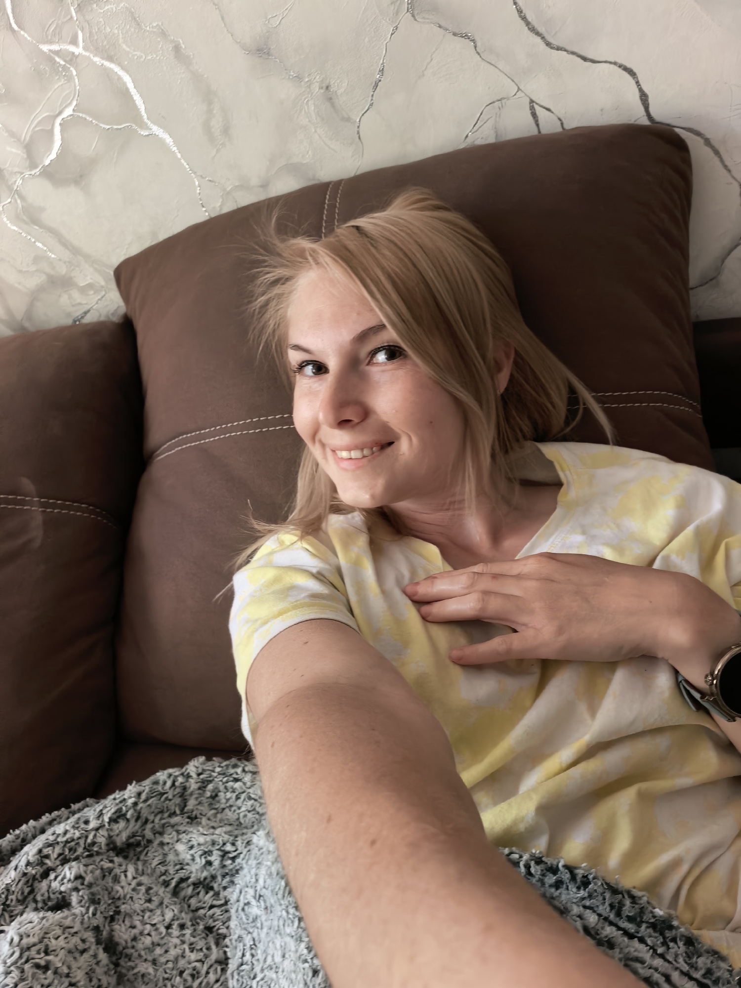 A light-skinned woman with blonde hair is lying on a brown couch or bed with a brown pillow. She is wearing a white and yellow tie-dye shirt and a watch. Her right hand is placed on her chest. The background includes a gray and white marbled wall. A gray blanket is on the couch. No location in Sterlitamak, Russia can be determined from the image.