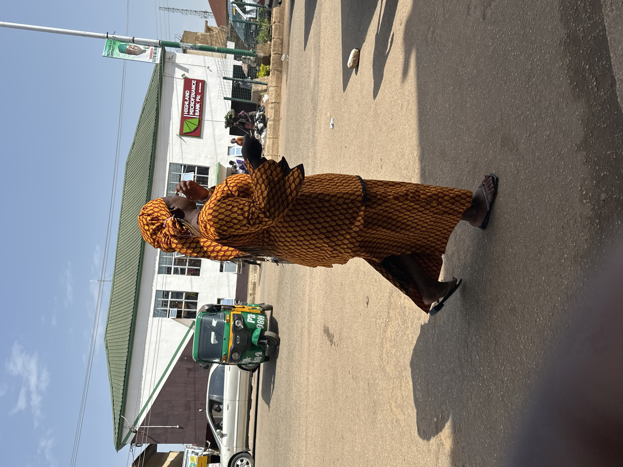 A woman in a patterned orange and yellow dress and headwrap is walking across a paved road in Bukuru, Nigeria. She is holding a black object to her mouth. A green and yellow auto-rickshaw is stopped behind her, and a silver sedan is in the background. A building with a green roof and white and brown walls is visible behind the vehicles. A banner displaying a picture of a man is on a pole to the right of the building. There are other people and vehicles in the distance. The sky is blue with a few scattered clouds.