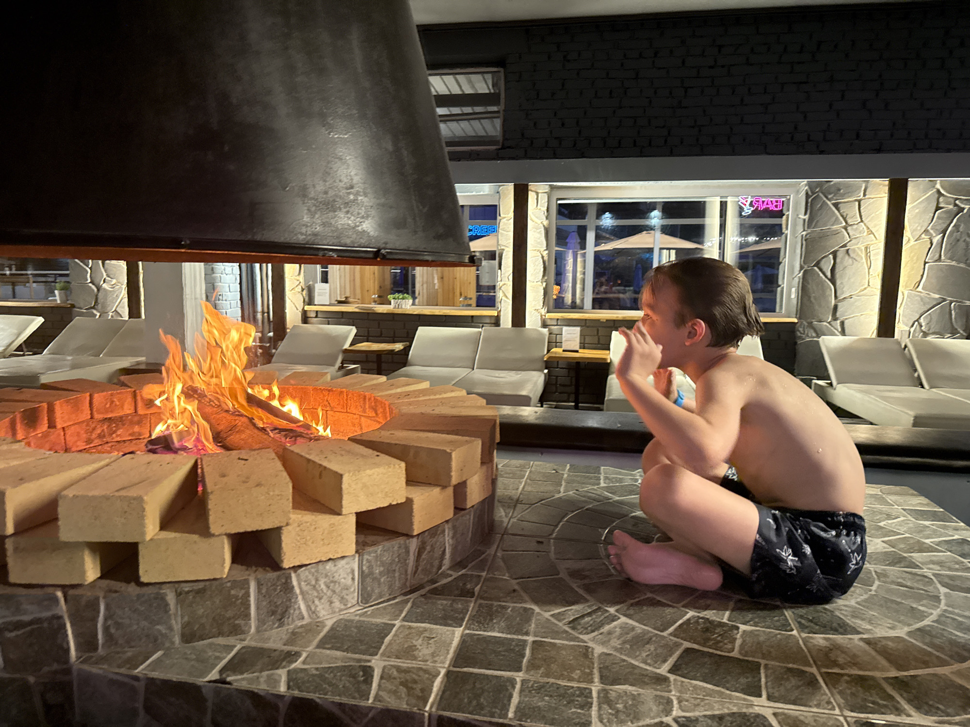 A shirtless, fair-skinned child sits cross-legged on a gray stone-tiled surface, their arms folded in contemplation near a brick fire pit ablaze with flames. The pit is positioned under a dark metal hood. Behind the child, lounge chairs are visible through a window, suggesting an outdoor patio setting, lit up with night lights. The background suggests an urban location with lit up signs and outdoor furniture. No location context about the city of Rostov-on-Don, Russia is evident in this image.